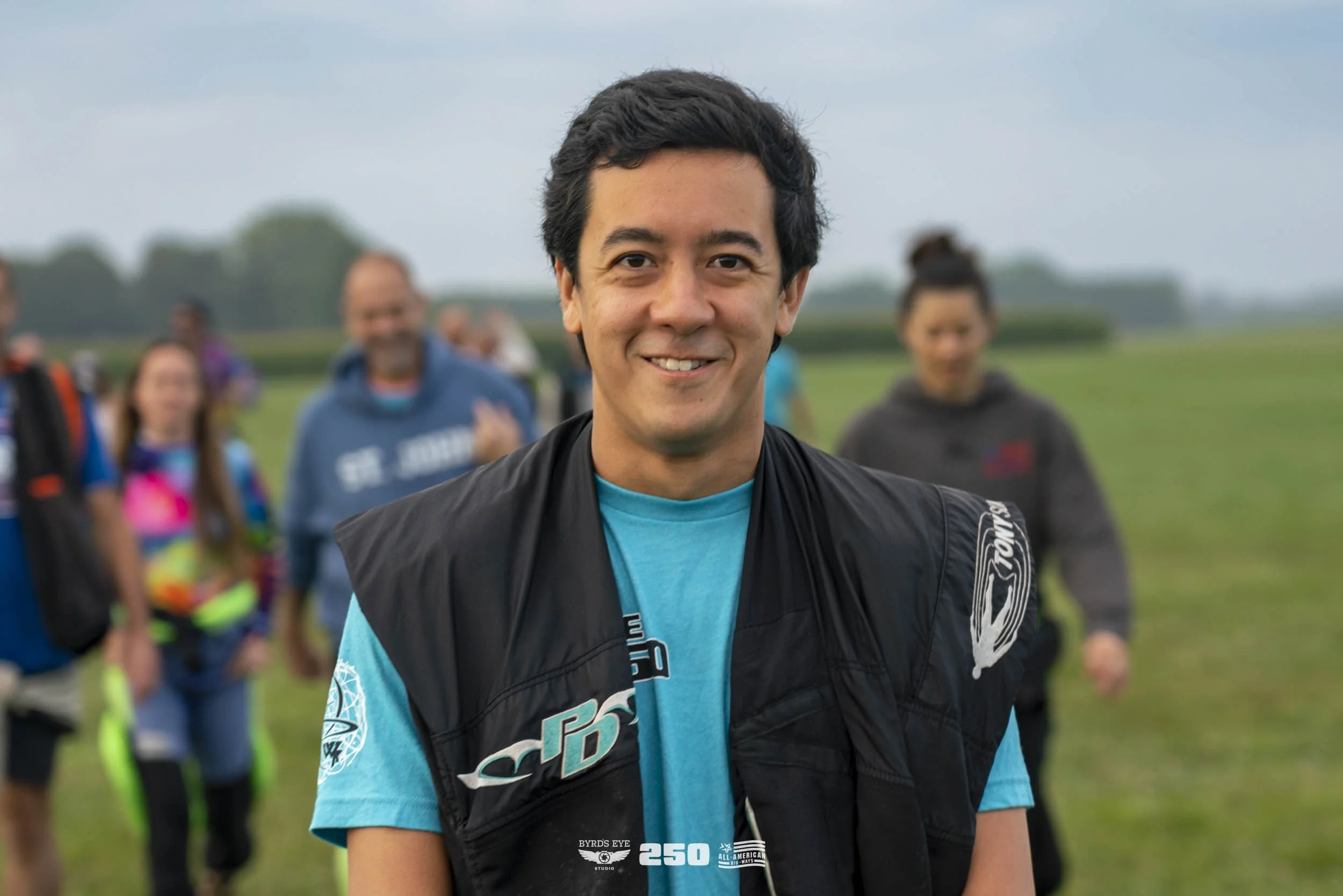 A young man smiling outdoors, wearing a blue shirt and black vest, with a group of people blurred in the background.