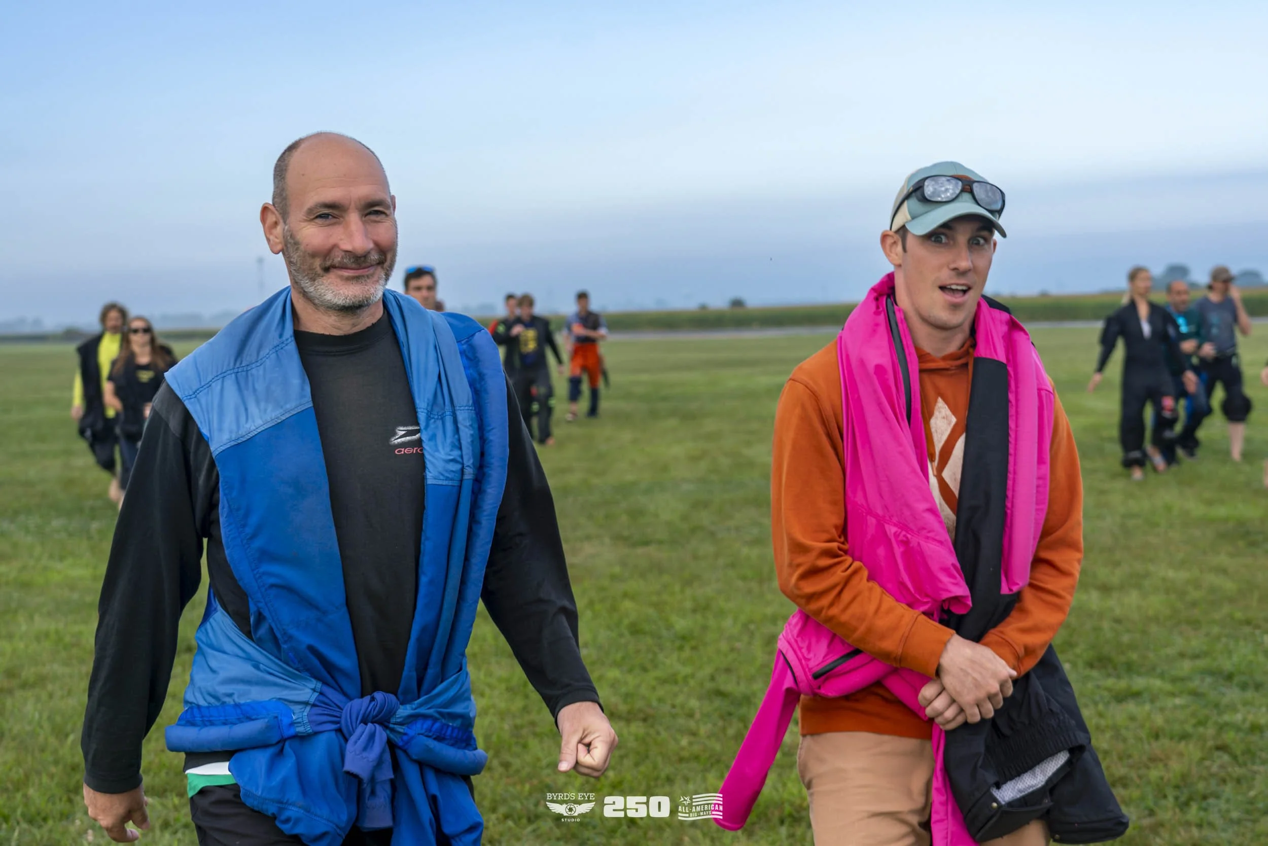A group of people walking outdoors on a grassy field, with two men in the foreground smiling and wearing colorful jackets, one in blue and the other in orange and pink. Several other people are visible walking in the background.