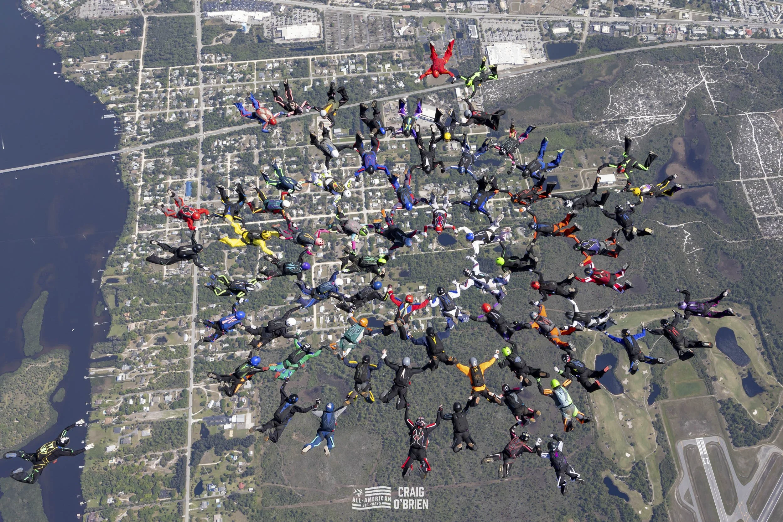 Multiple skydivers in various colored jumpsuits and helmets free-falling over a landscape with water, roads, and buildings, holding hands in a circular formation.