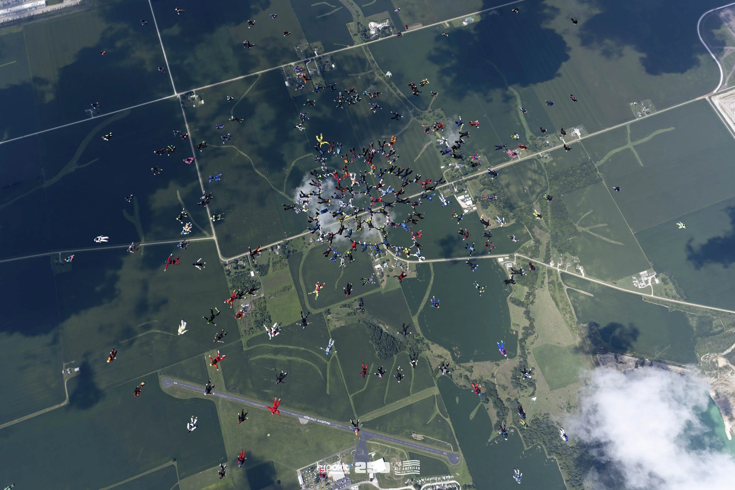 A large group of skydivers forming a pattern in the sky over a green landscape with lakes, fields, and roads.