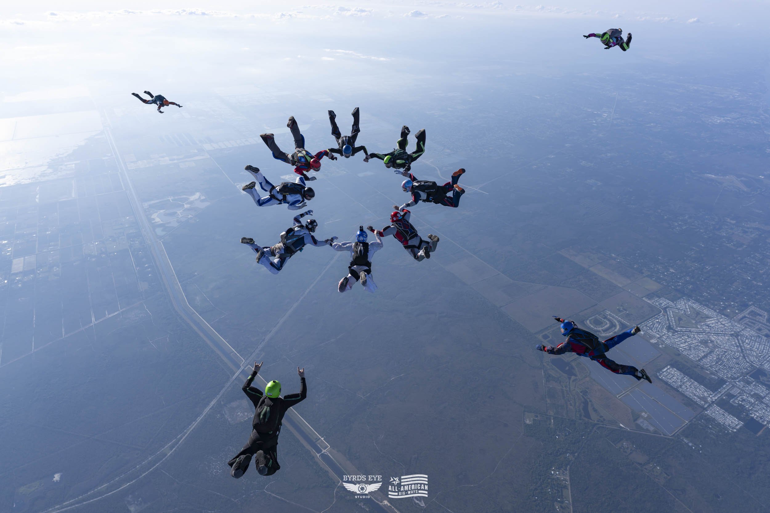 Group of skydivers in free fall, holding hands in a circle, with others scattered around, high above a landscape of fields and roads.