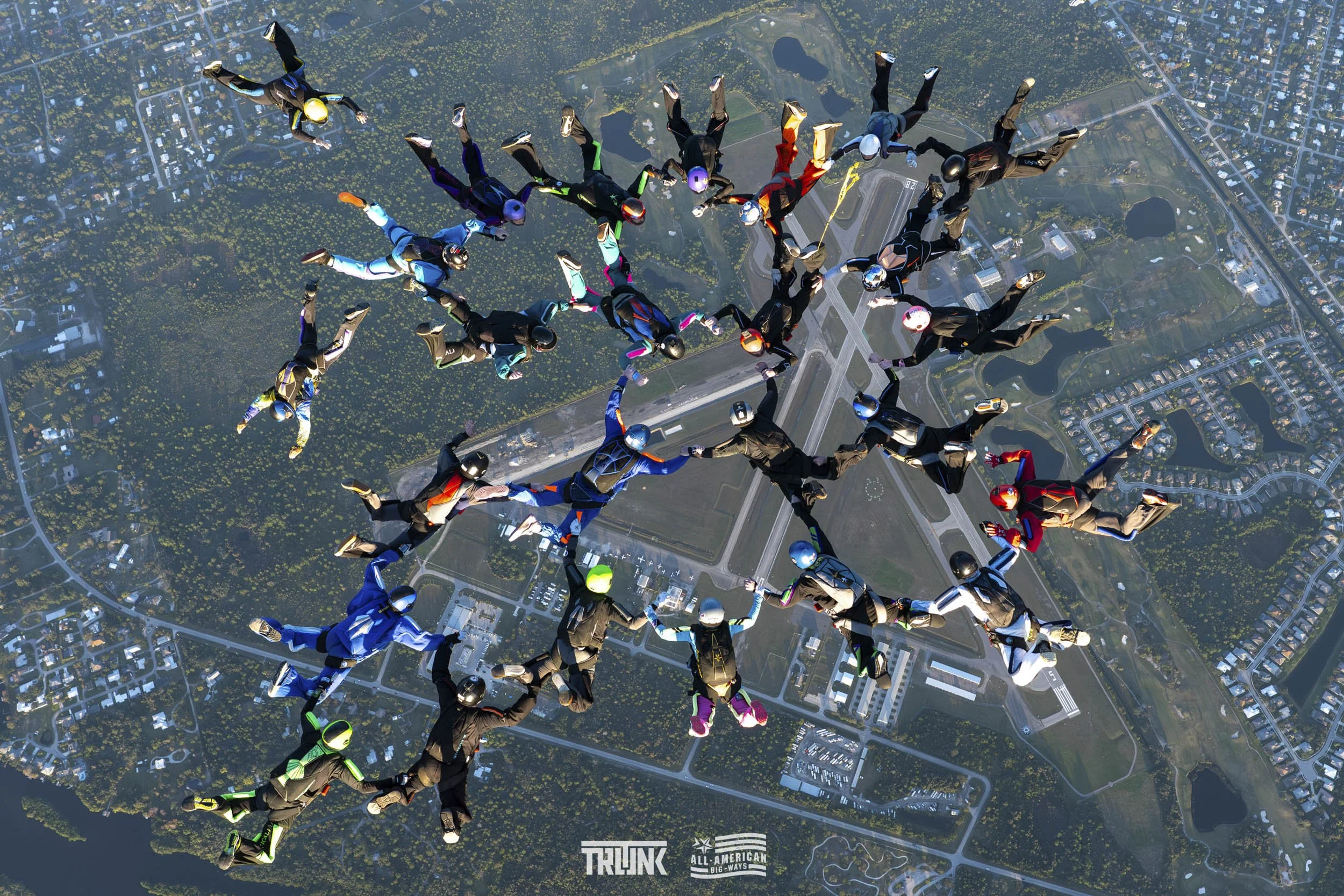 A group of skydivers holding hands in a circle during freefall over a cityscape with buildings, roads, and greenery.