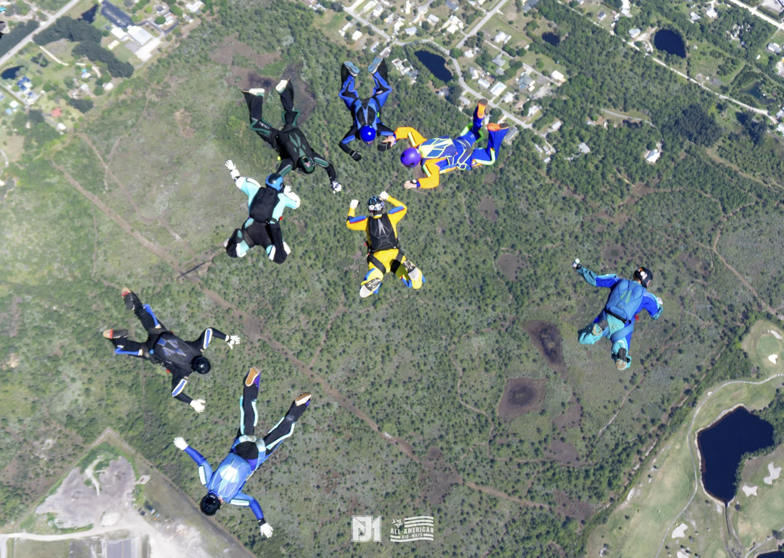 Group of seven skydivers in colorful jumpsuits free-falling over a green landscape with trees, fields, a lake, and some buildings visible below.