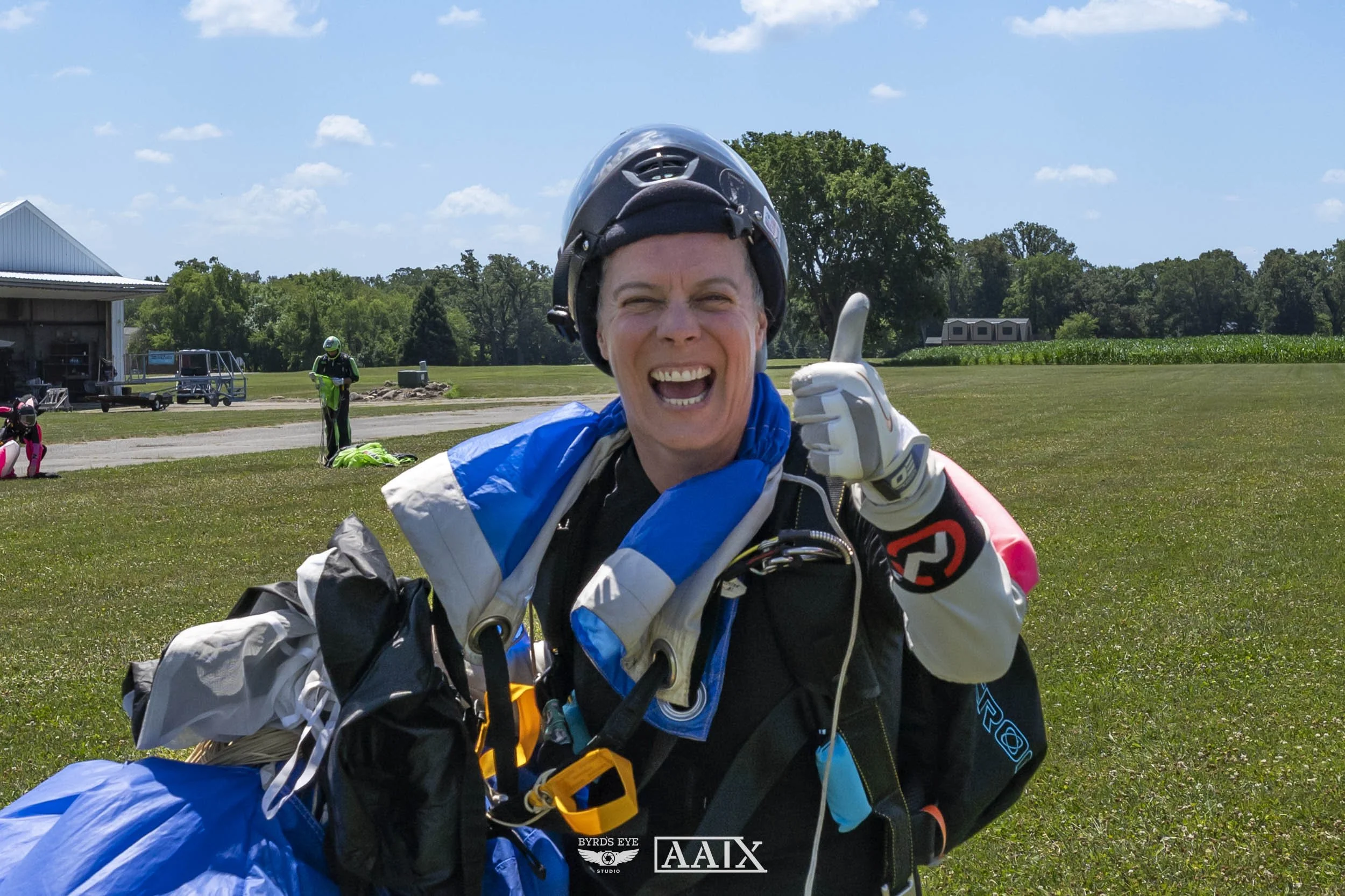 A woman in skydiving gear smiling and giving a thumbs-up outdoors on a grassy field, with a clear blue sky and trees in the background.