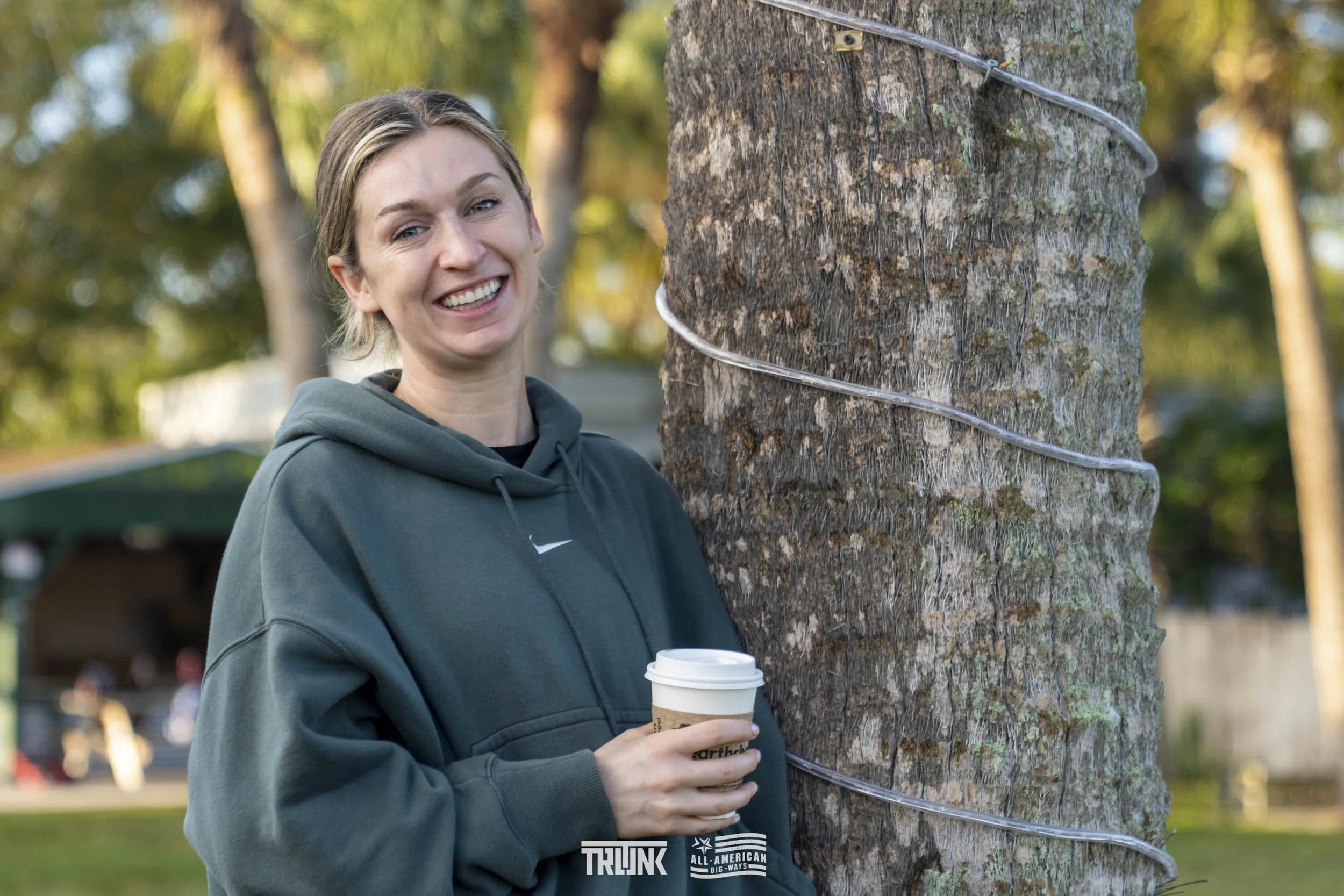 A woman smiling outdoors holding a coffee cup, standing next to a tree wrapped with metal wires.