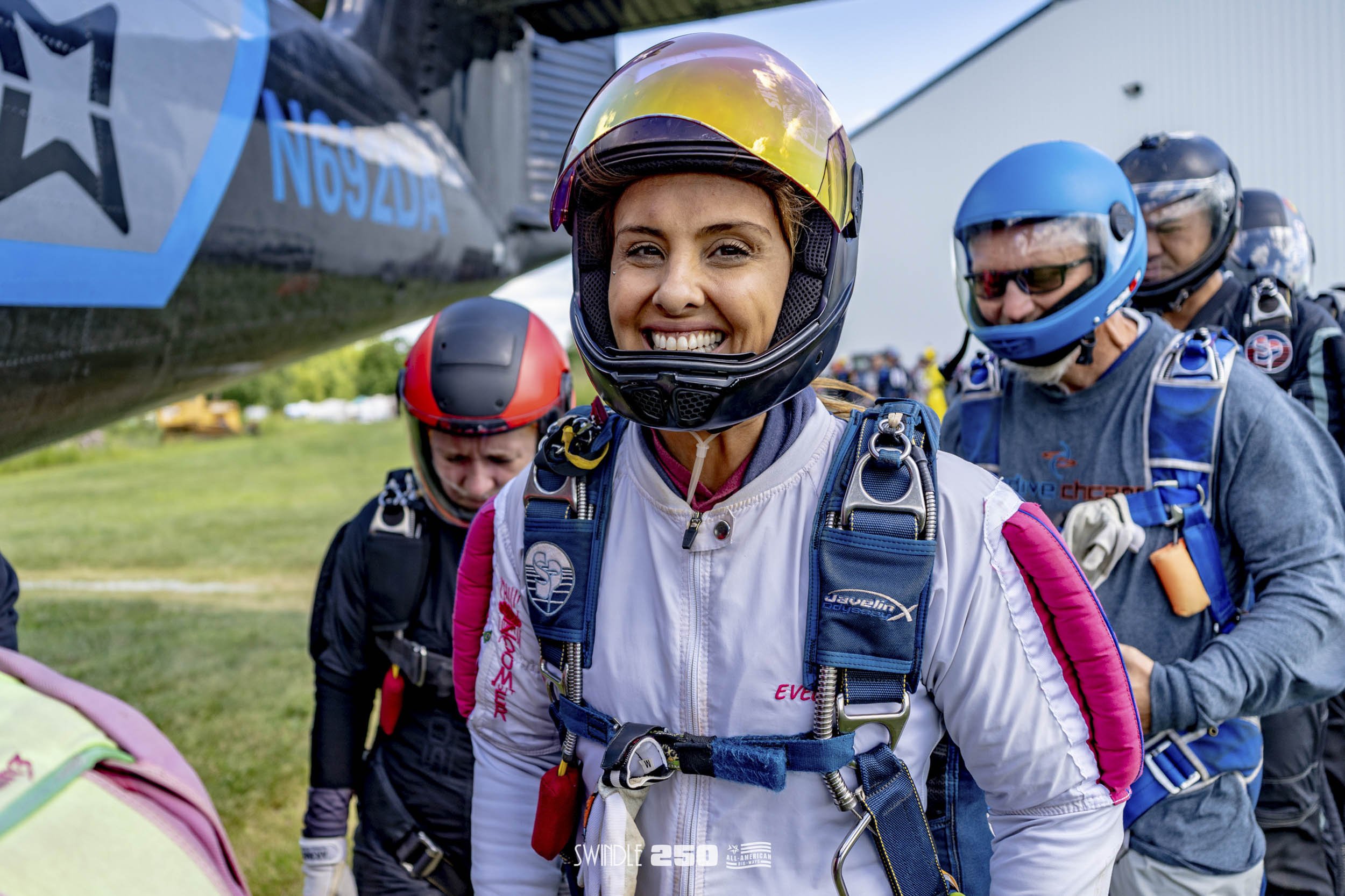 A group of skydivers in jumpsuits and helmets standing outdoors near a helicopter, preparing for a jump. The woman in front is smiling.