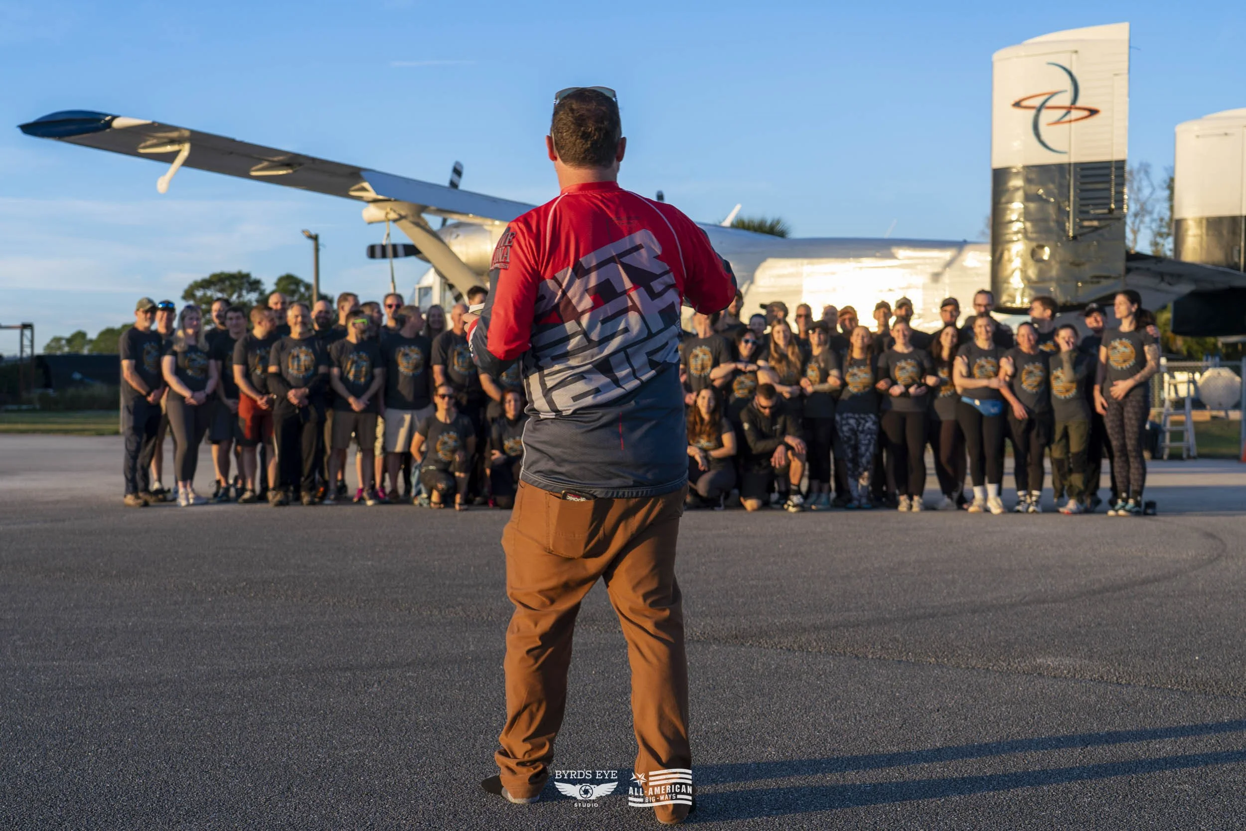 Man standing on tarmac facing a large group of people with an aircraft in the background during sunset.