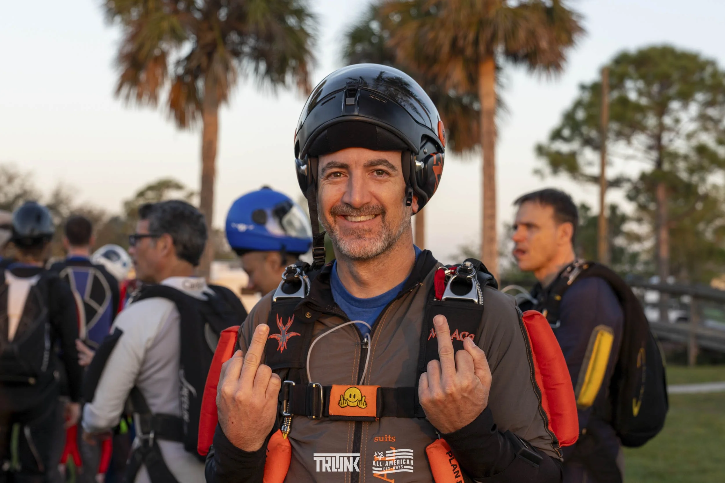 A smiling man wearing a motorcycle helmet and gear, showing middle fingers, at an outdoor gathering with palm trees and other people in the background at sunset.