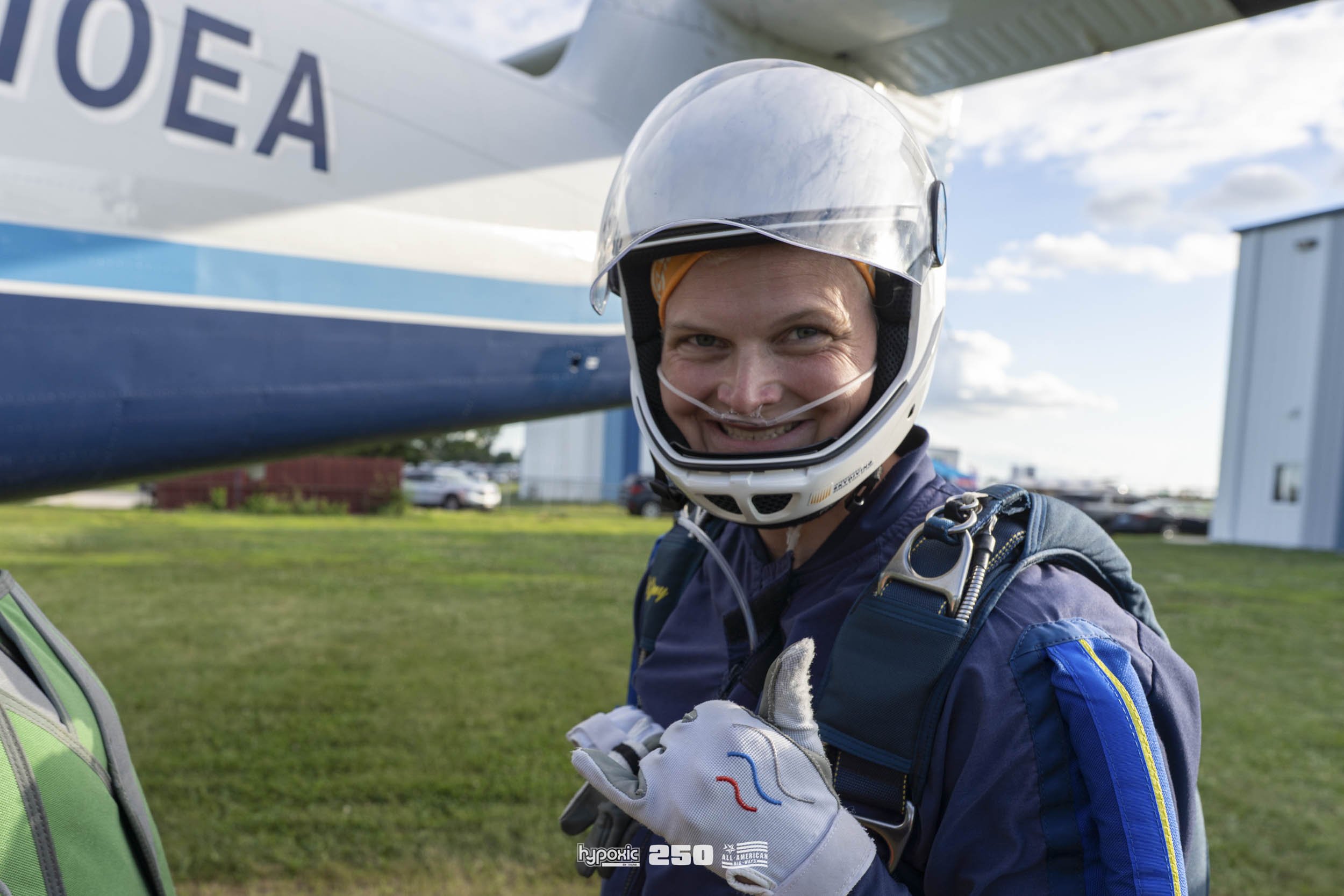 A smiling man in a helmet and skydiving suit standing next to a small aircraft, giving a thumbs up before skydiving outdoors on a cloudy day.