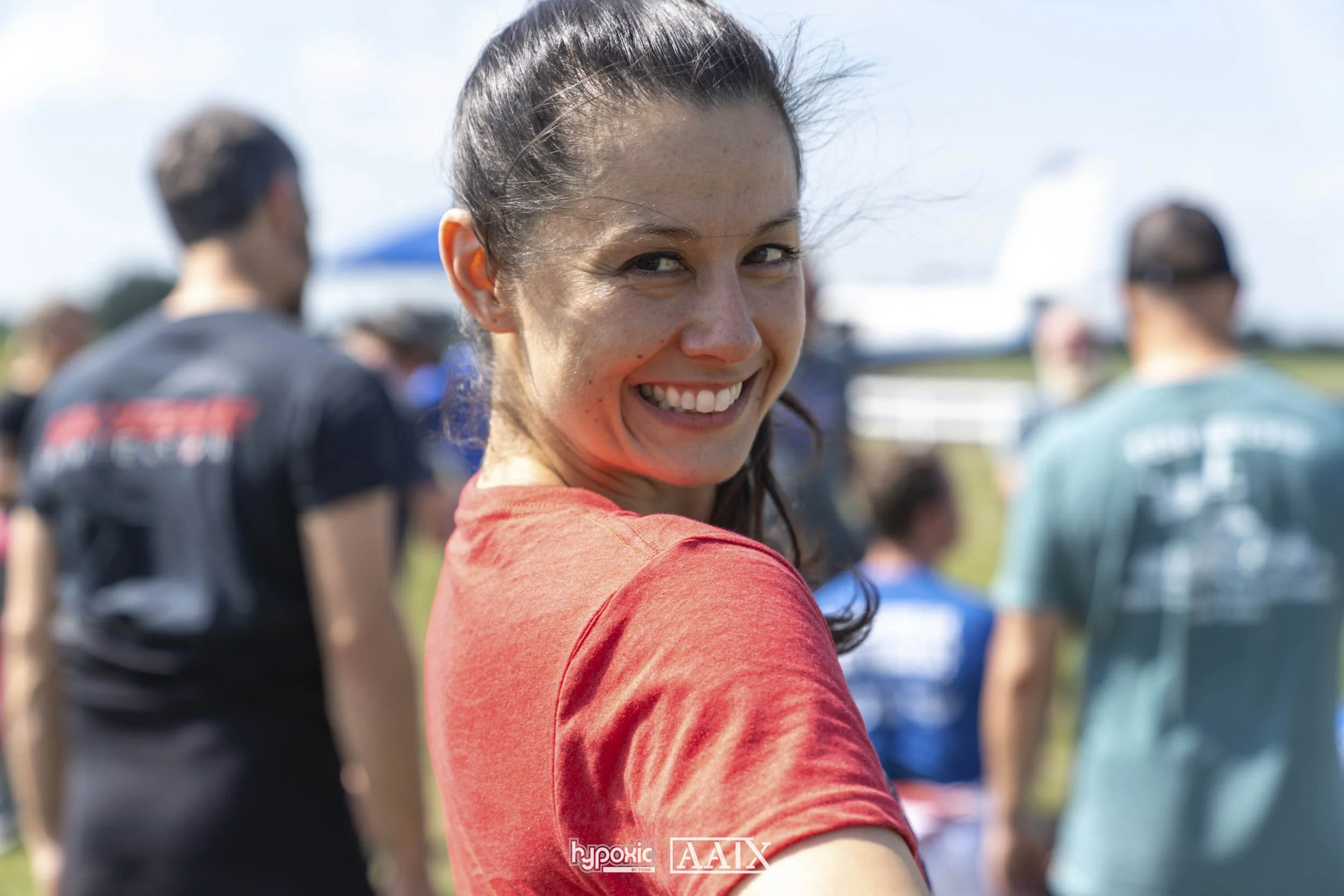 A woman in a red shirt smiling at the camera with people in the background at an outdoor event.