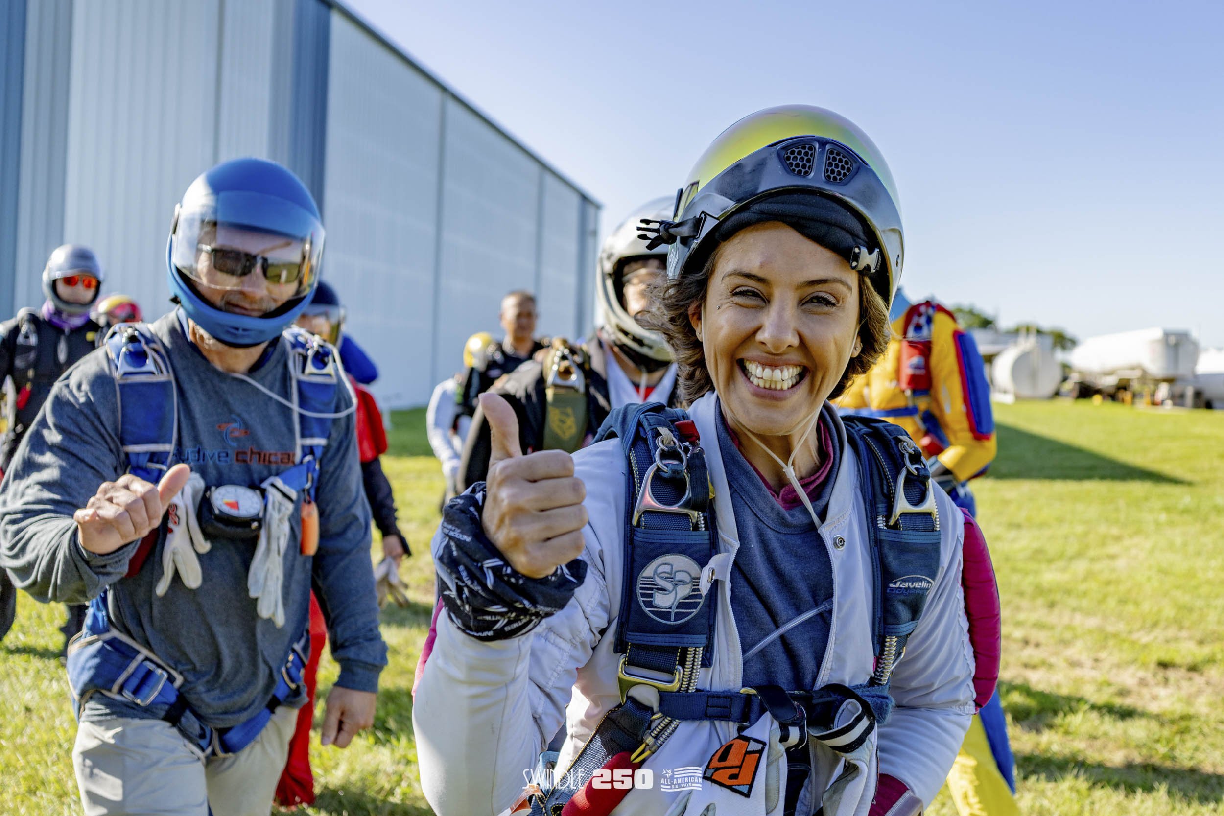 Group of skydivers preparing for jump, with smiling woman in foreground giving a thumbs-up and wearing a helmet, others in helmets and jump gear in the background, outdoor setting with clear blue sky.