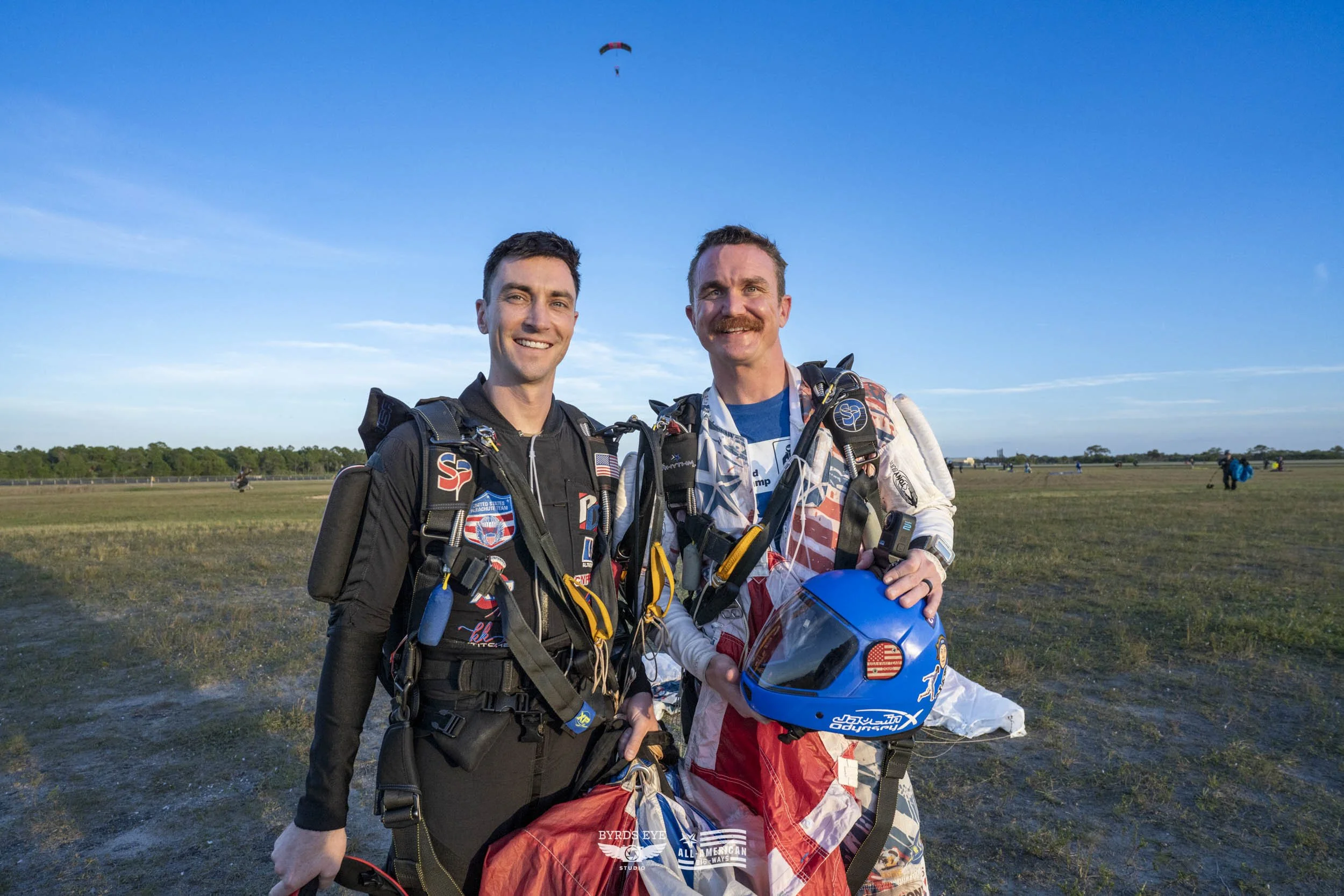 Two skydivers in jumpsuits with parachutes pose together on an open field, with a blue sky and a parachute descending in the distance.