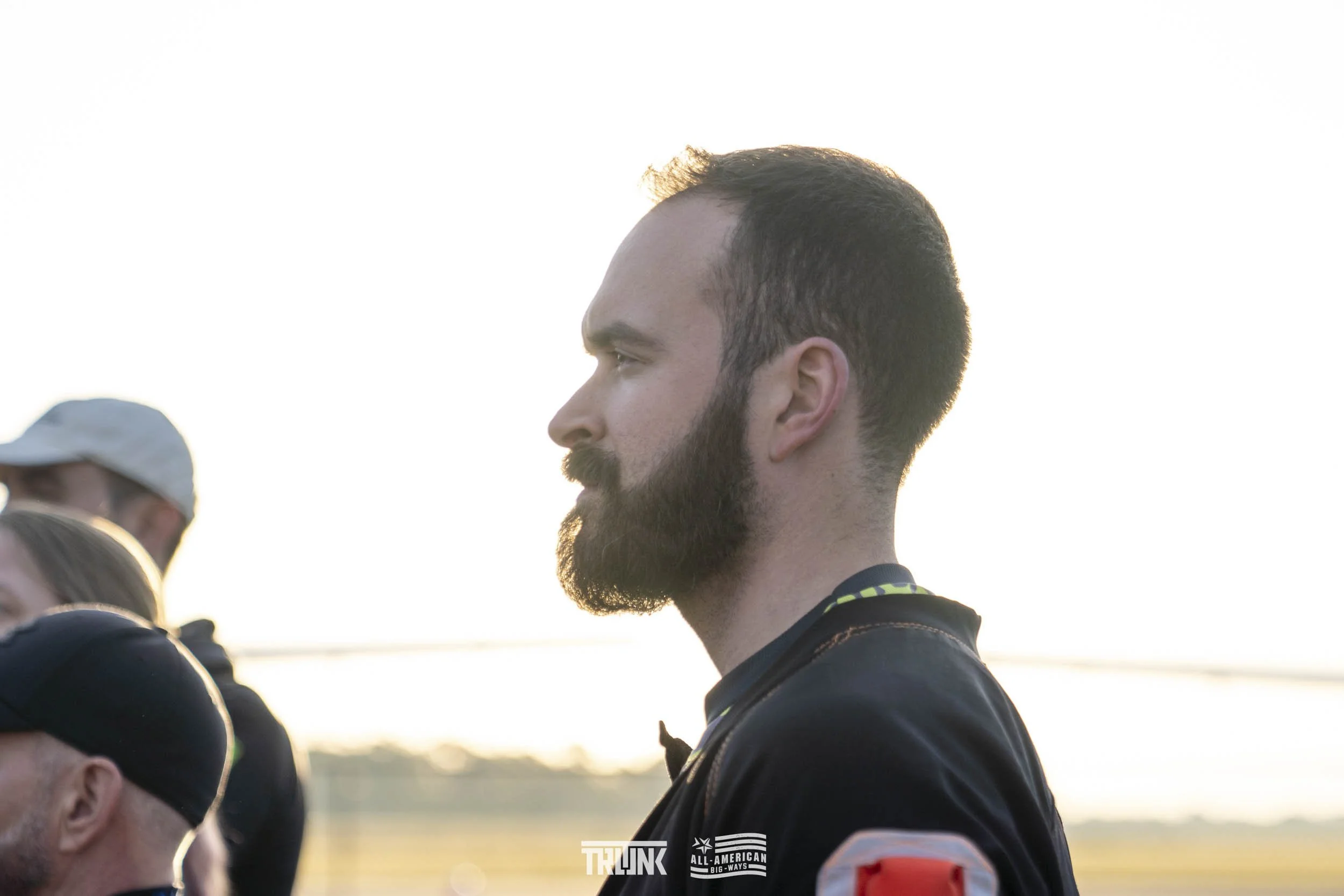 A man with a beard and short dark hair stands outdoors in profile view during sunset. Several other people are in the background, some wearing hats.