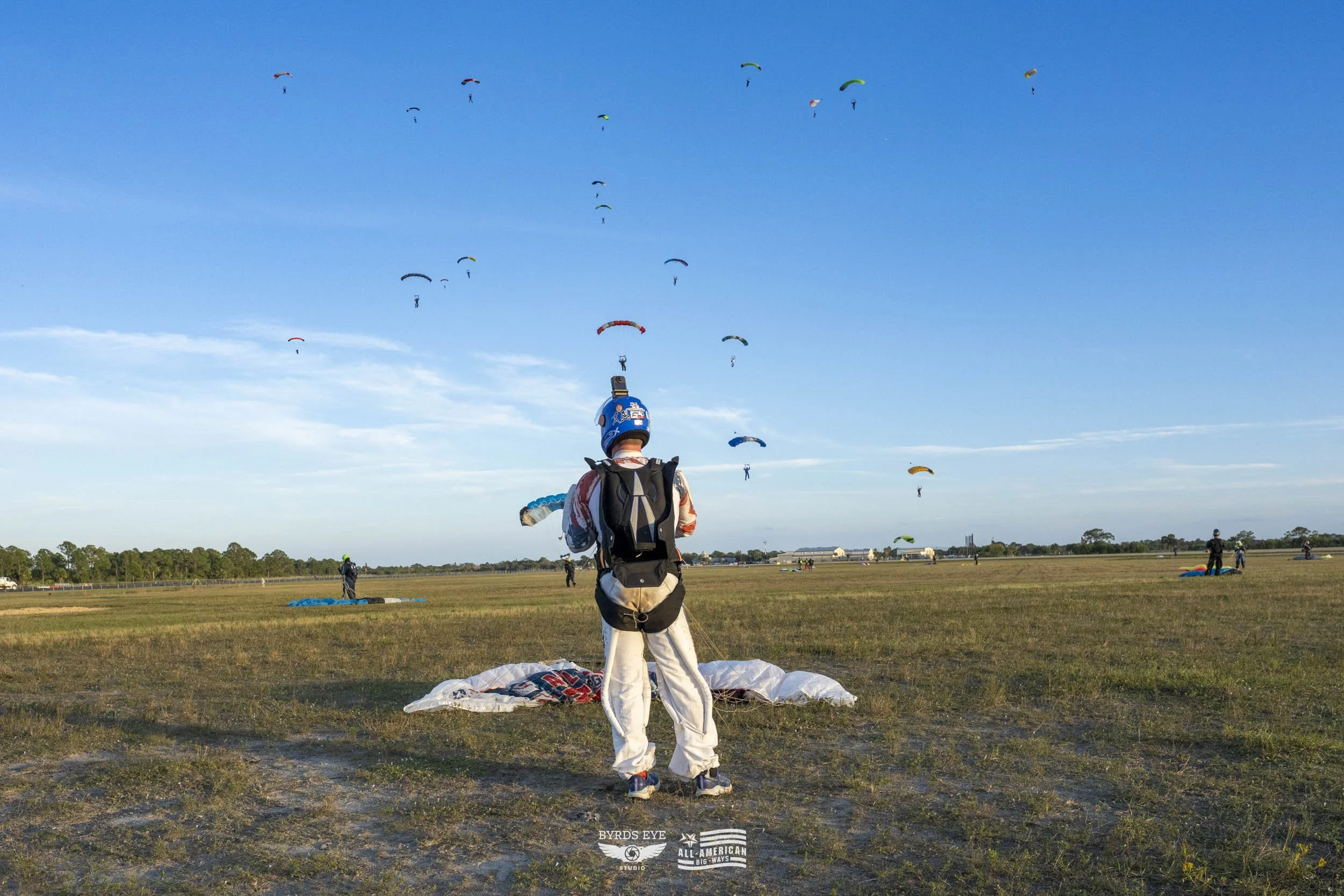 A person standing on a grassy field wearing a helmet and backpack, preparing for a paragliding jump. Several paragliders are airborne in the blue sky above.