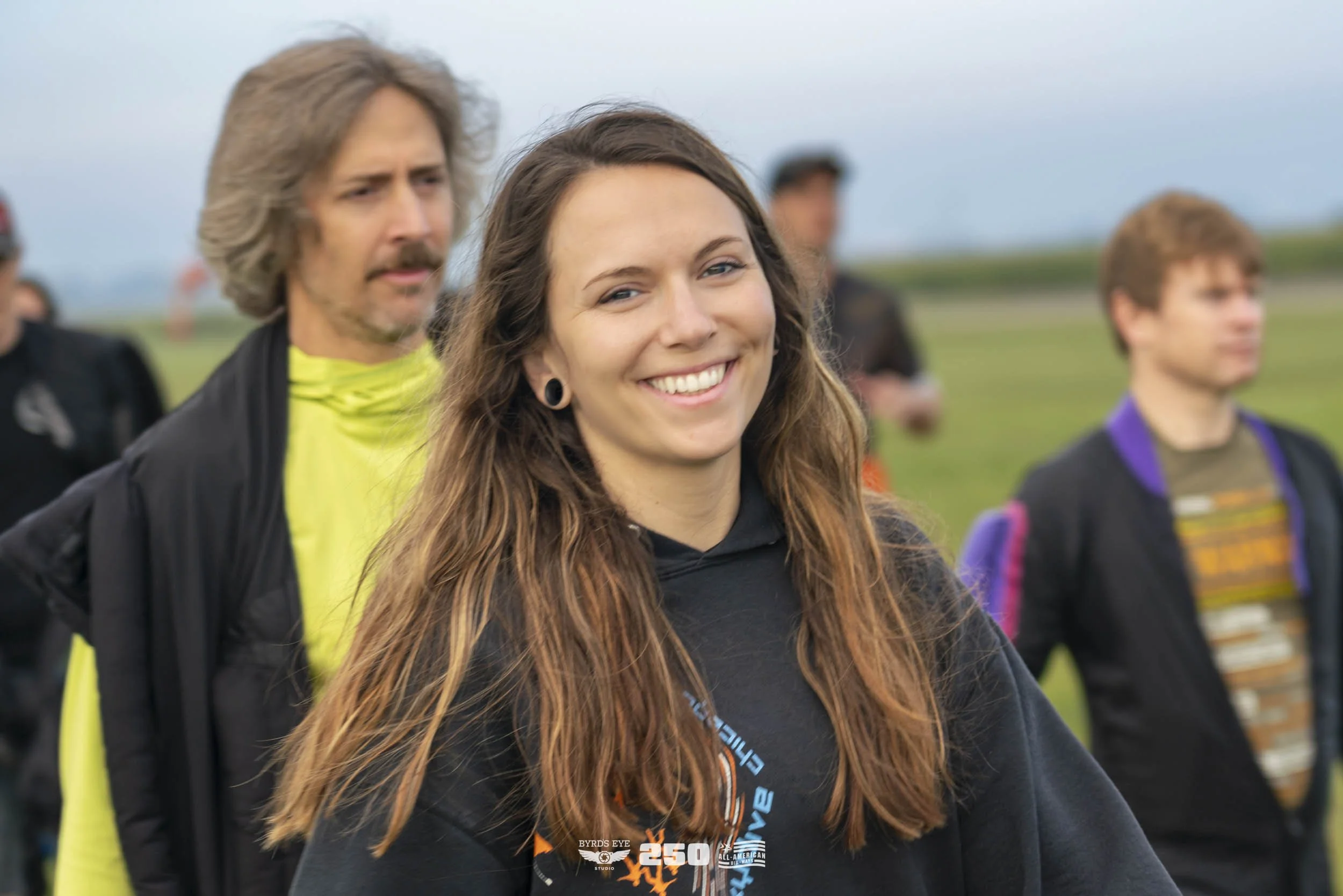 A group of people outdoors, with a young woman smiling in the foreground, wearing a black hoodie, and others blurred in the background on a cloudy day.