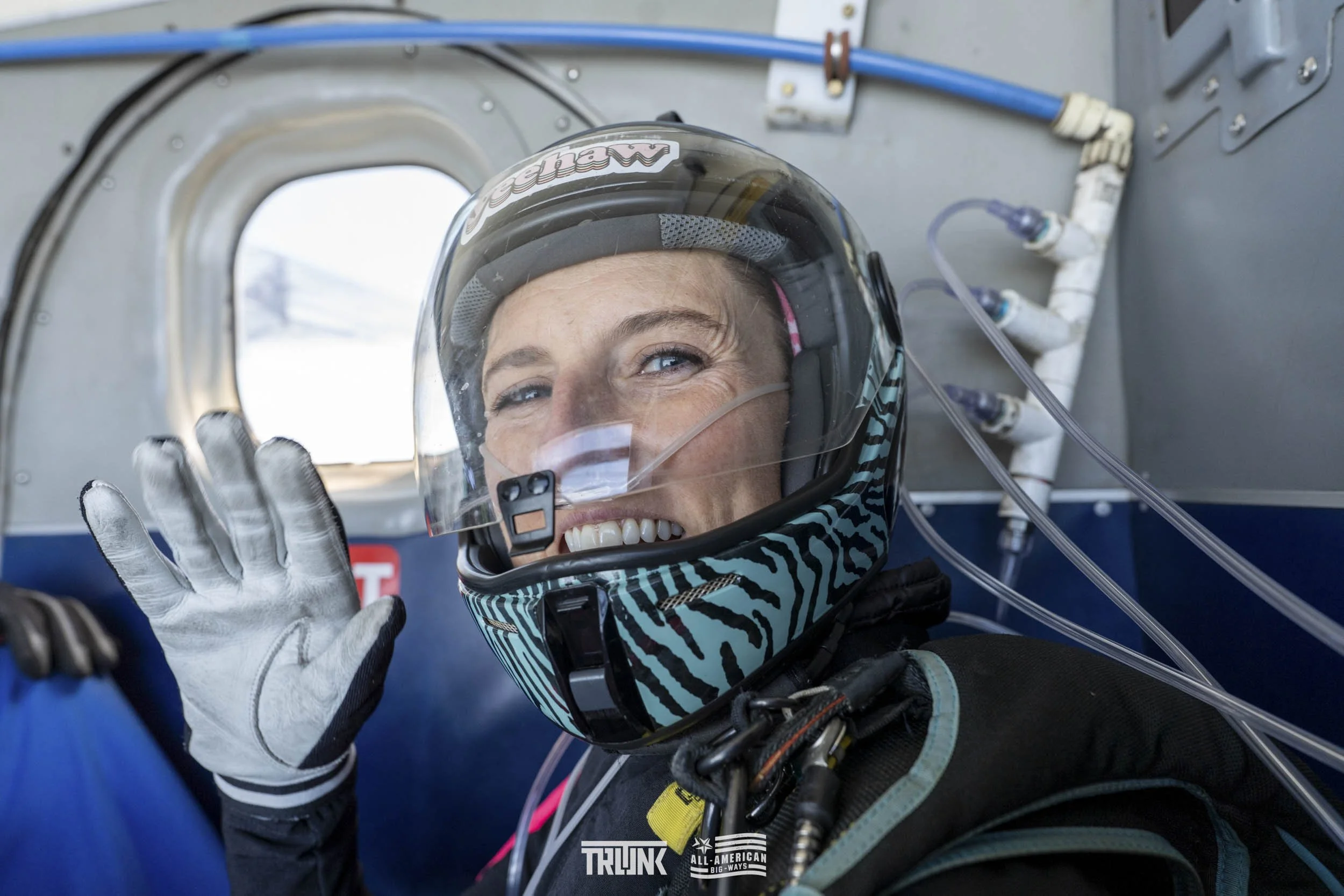 Close-up of a smiling female race car driver wearing a helmet and gloves, inside a race car with equipment and tubing visible in the background.