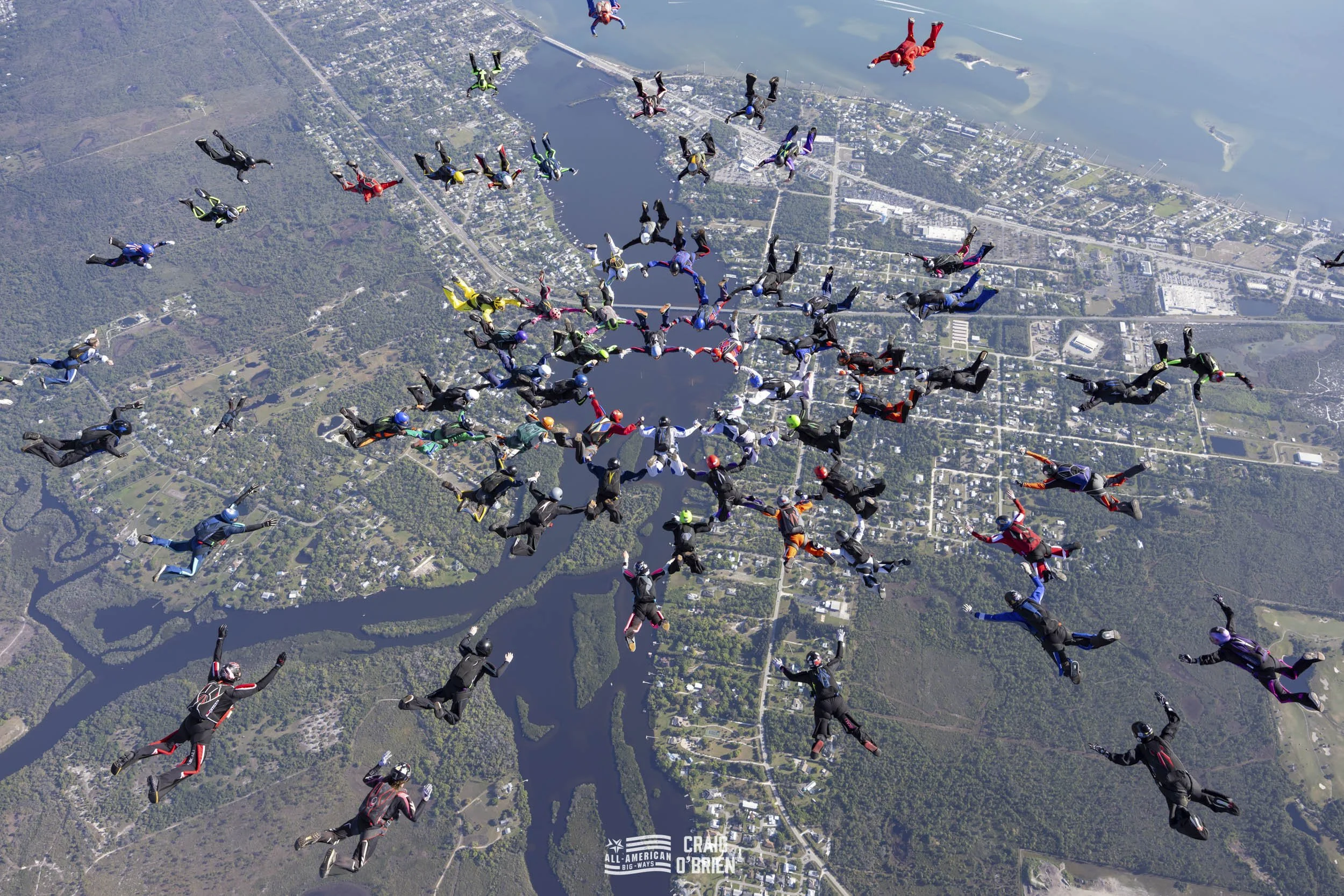 A large group of skydivers wearing colorful jumpsuits and helmets, forming a pattern while freefalling over an aerial view of a landscape with water bodies, roads, and green areas.