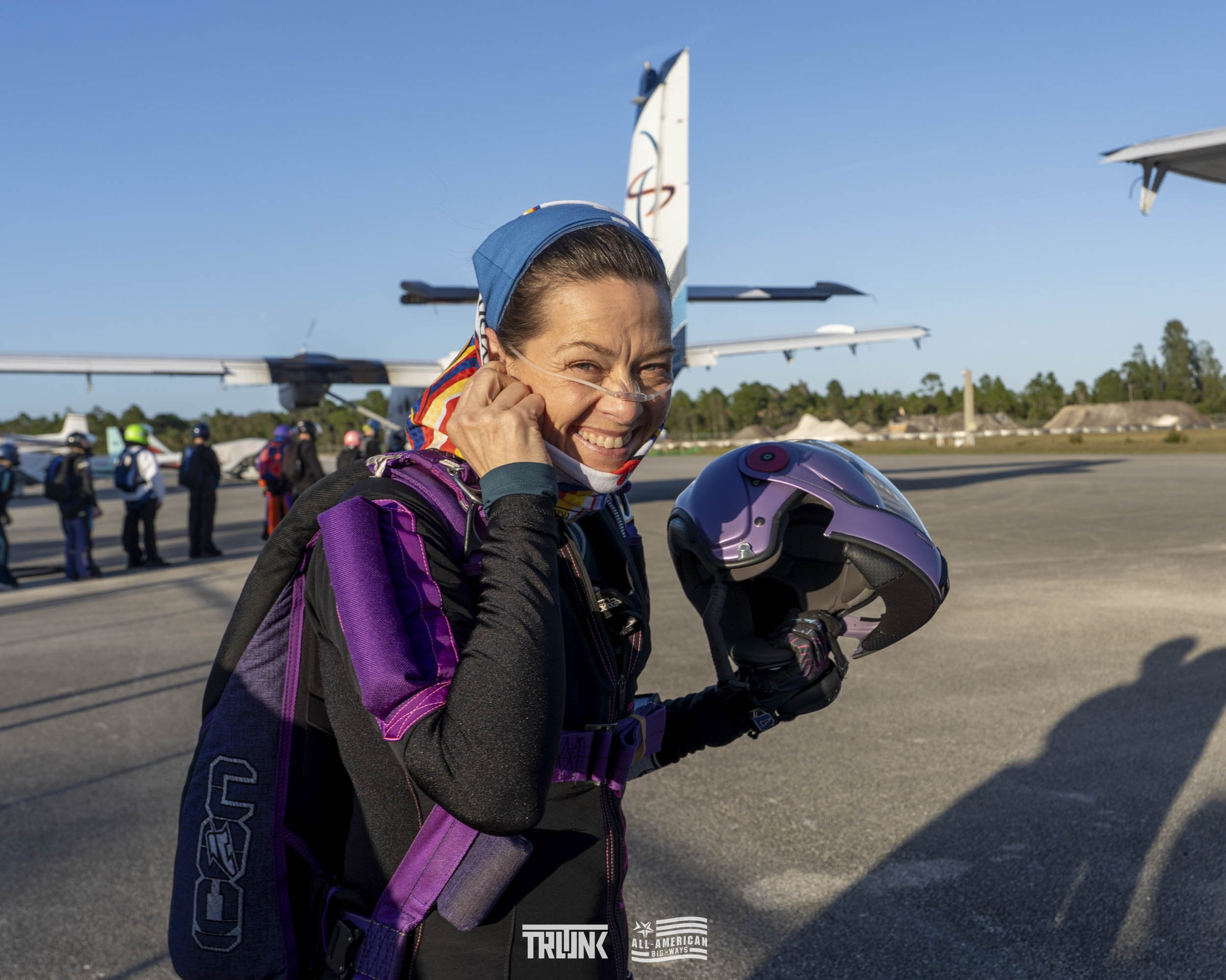 A smiling woman in an aviation jumpsuit holding a purple helmet, standing on an airport tarmac with small aircraft in the background.