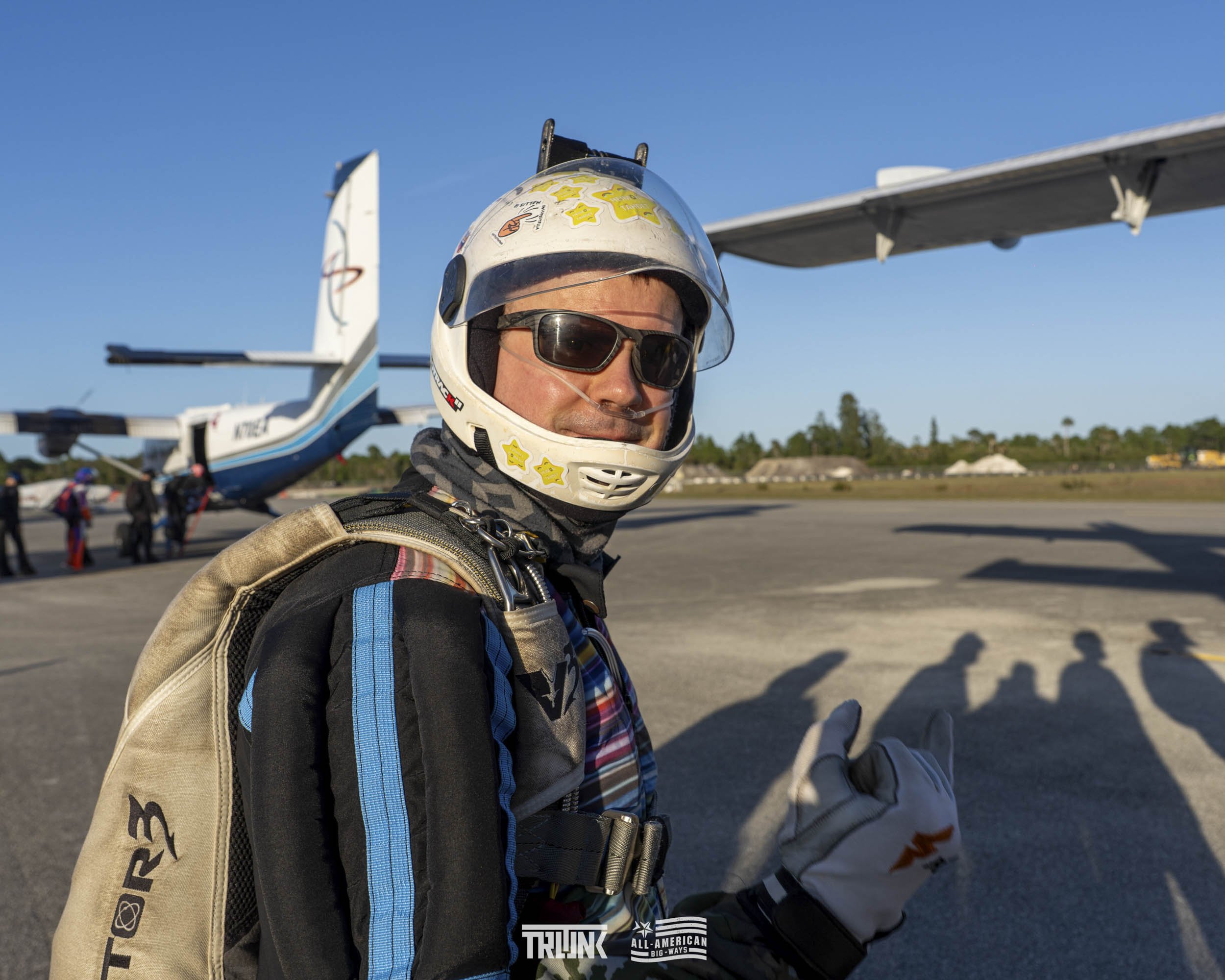 A man in racing gear, wearing a helmet and sunglasses, stands on an airstrip with a small airplane in the background.