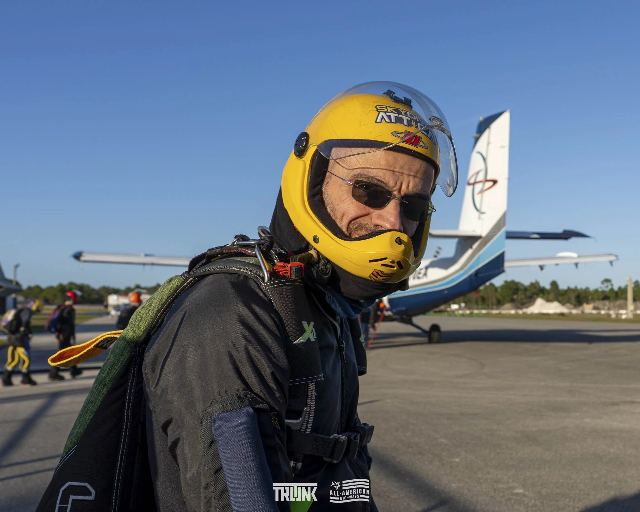 A man in a yellow skydiving helmet and sunglasses stands in front of a small airplane on an airstrip, preparing for a jump.