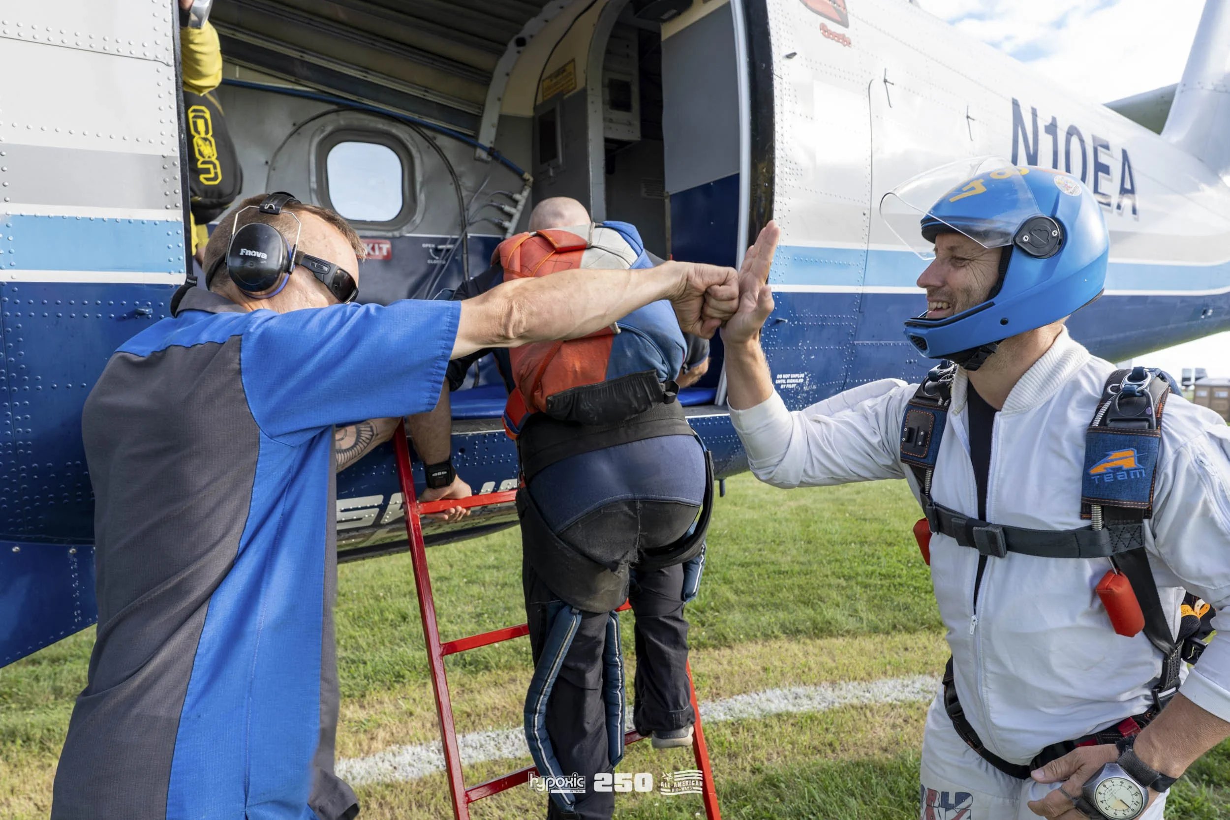 Skydiver in jumpsuit is climbing into a blue helicopter with assistance from instructors on the ground, who are giving high-fives, with grassy field in the background.