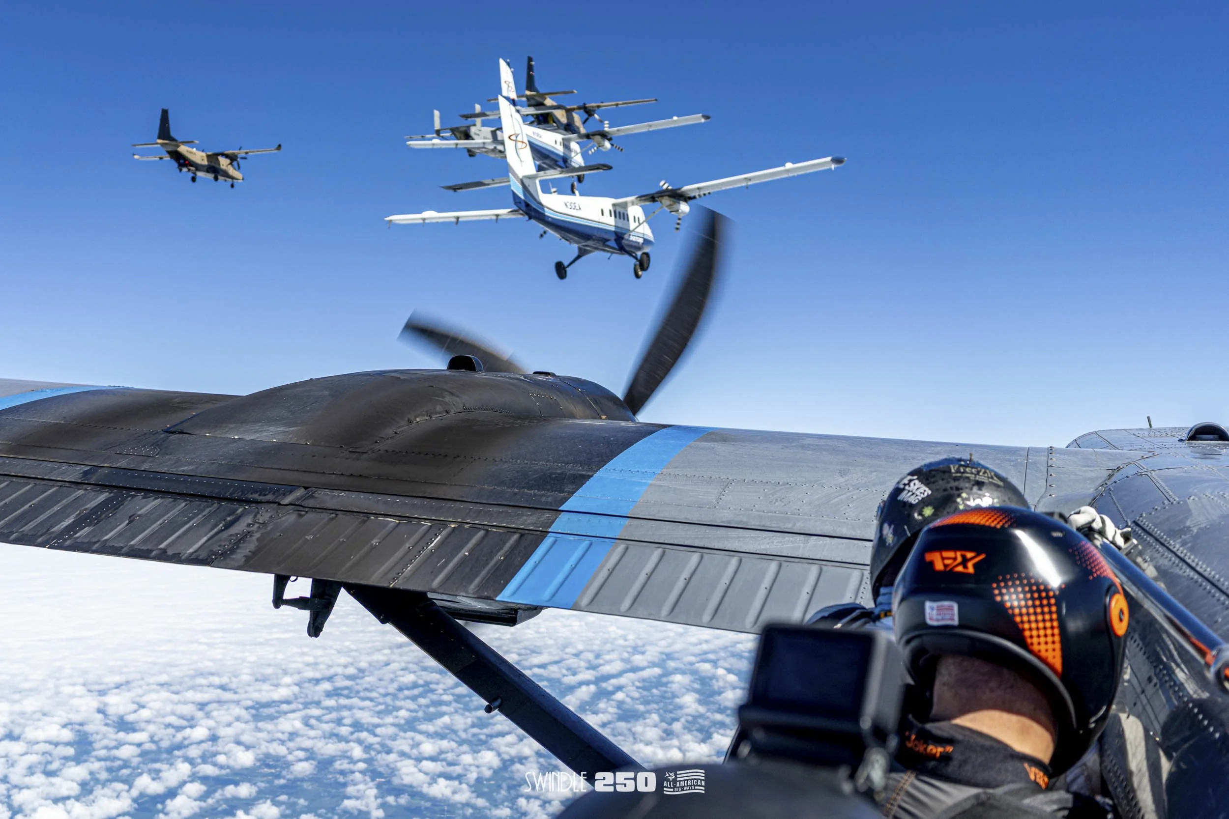 Aerial view of military aircraft flying in formation above the clouds, with a person wearing a helmet in the foreground taking a selfie with the planes in the background.