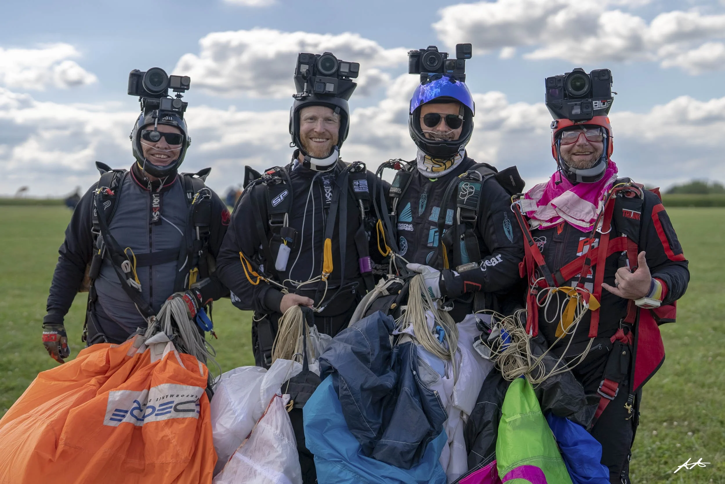 Four skydivers in jumpsuits and helmets with cameras on top, standing outdoors on a grassy field with clouds in the sky, holding parachutes.