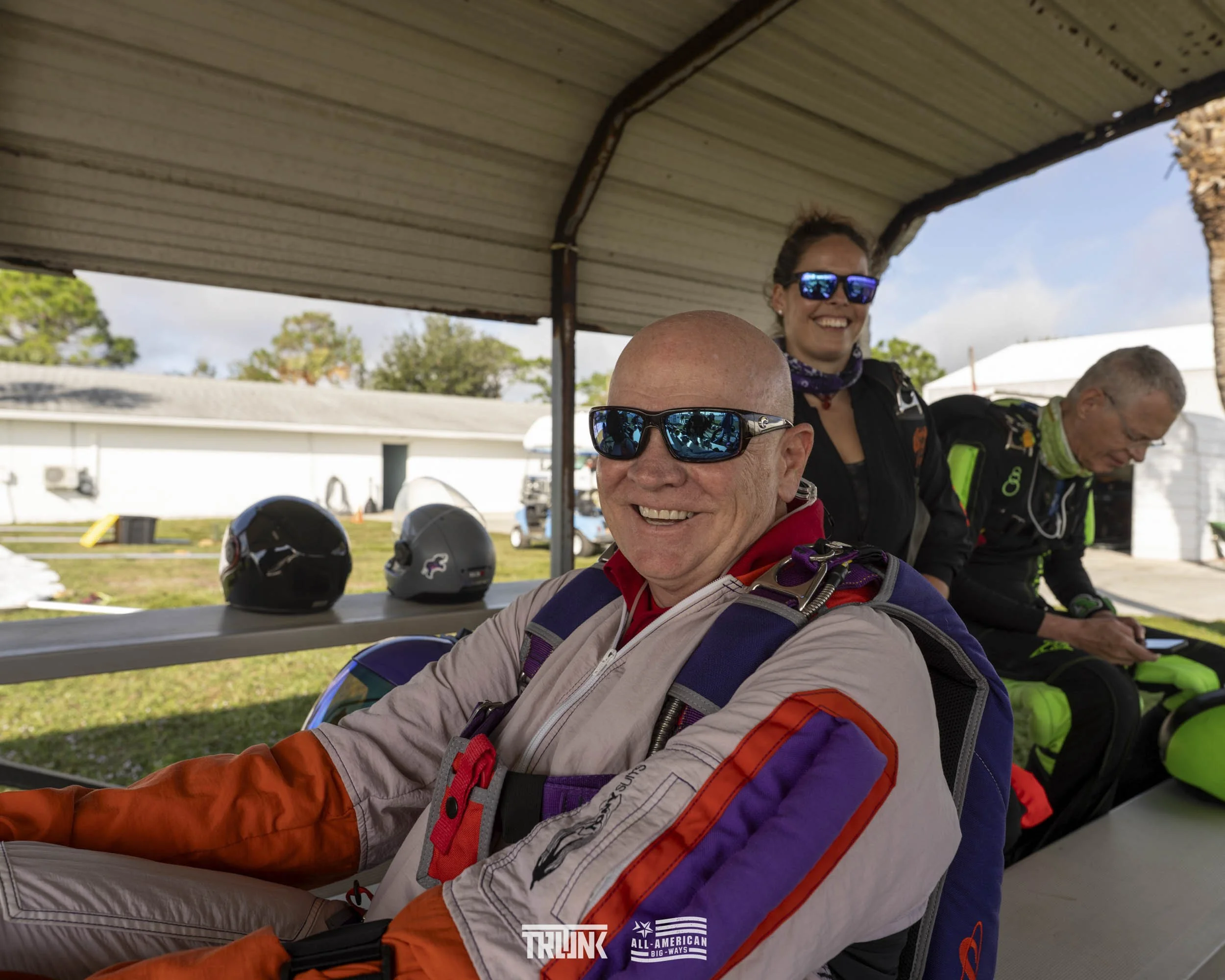 A group of skydivers at a jump site, with helmets and gear on a table behind them, one man smiling at the camera wearing sunglasses, a woman above him smiling, and another man looking at his phone, all under a metallic roof.