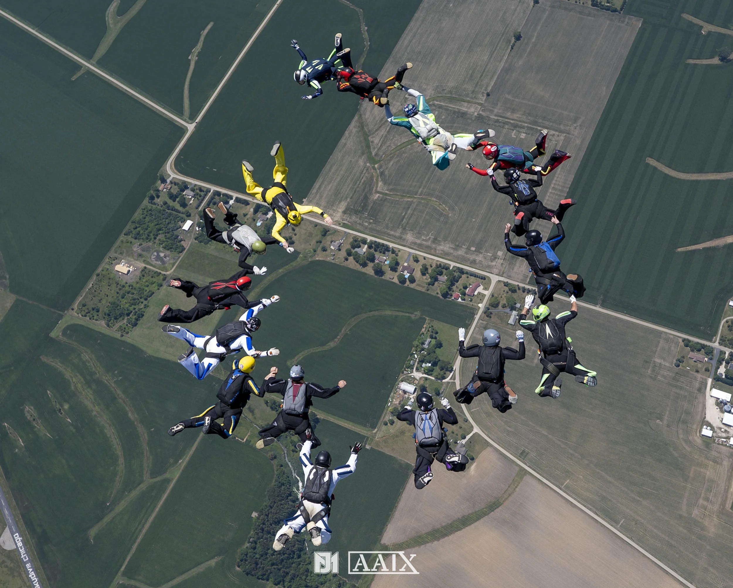 Group of skydivers in freefall formation over farmland.