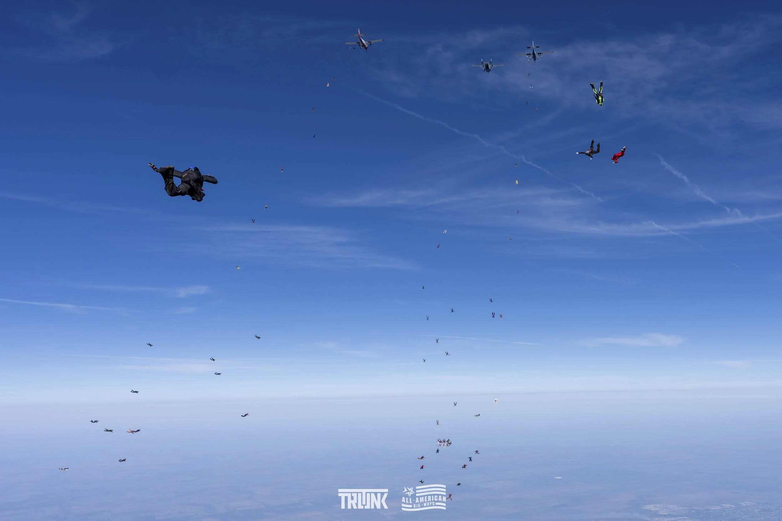 Skydivers in freefall formation with visible parachutes opening and falling through the blue sky, with some aircraft flying above.