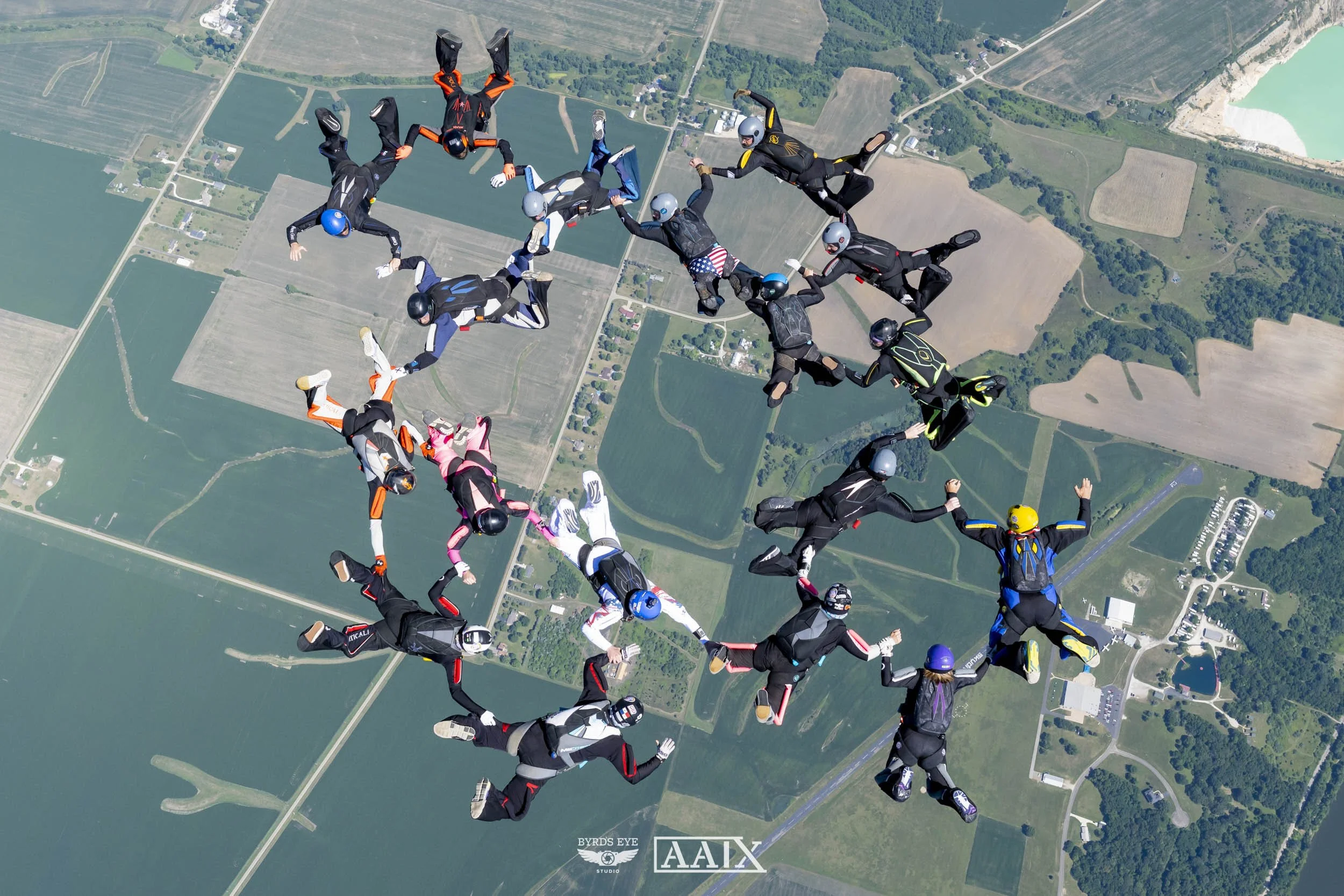 Group of skydivers forming a circle while floating over farmland, with colorful jumpsuits and helmets, holding hands in mid-air.
