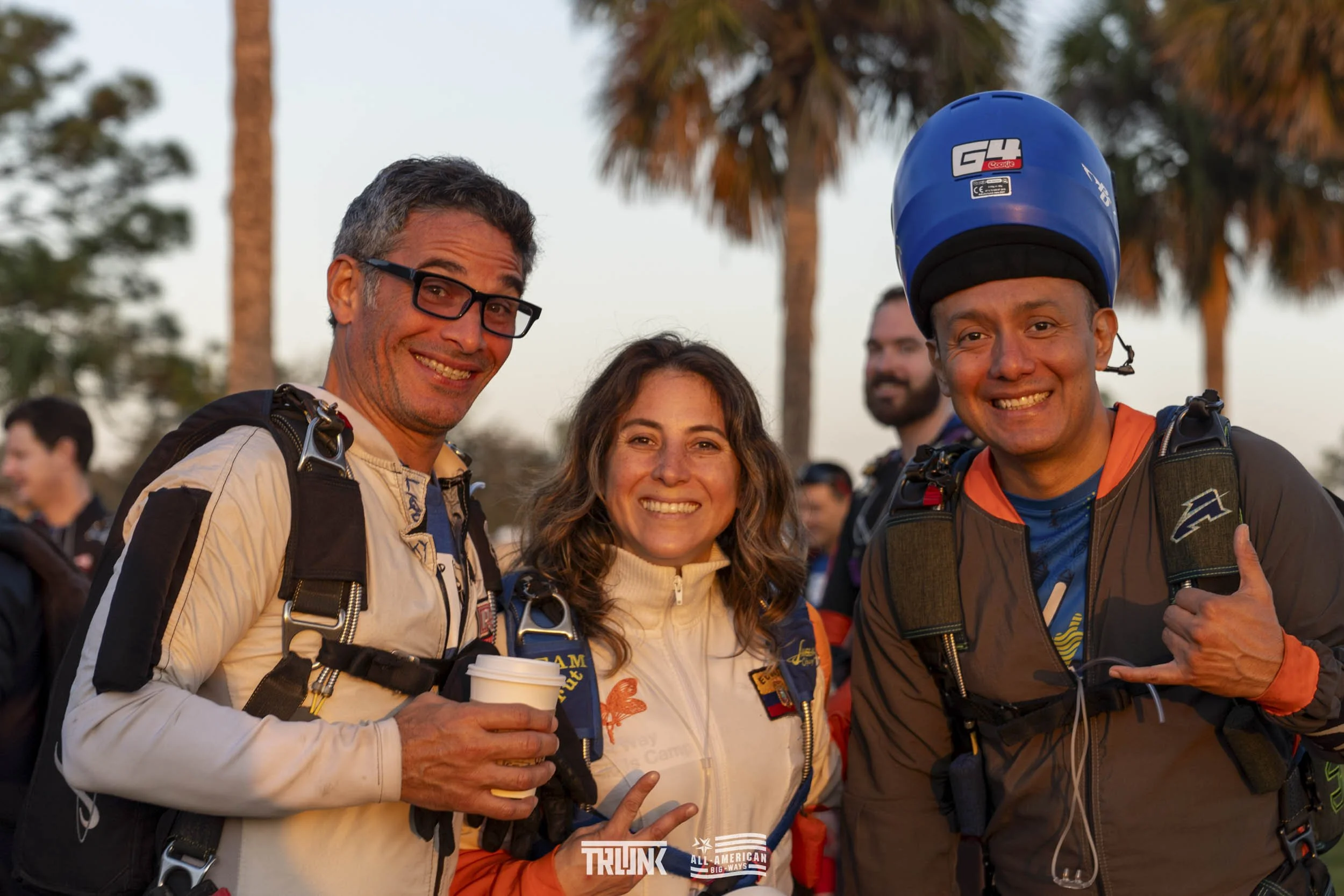 Three smiling skydivers in jumpsuits and helmets at an outdoor event with palm trees in the background.