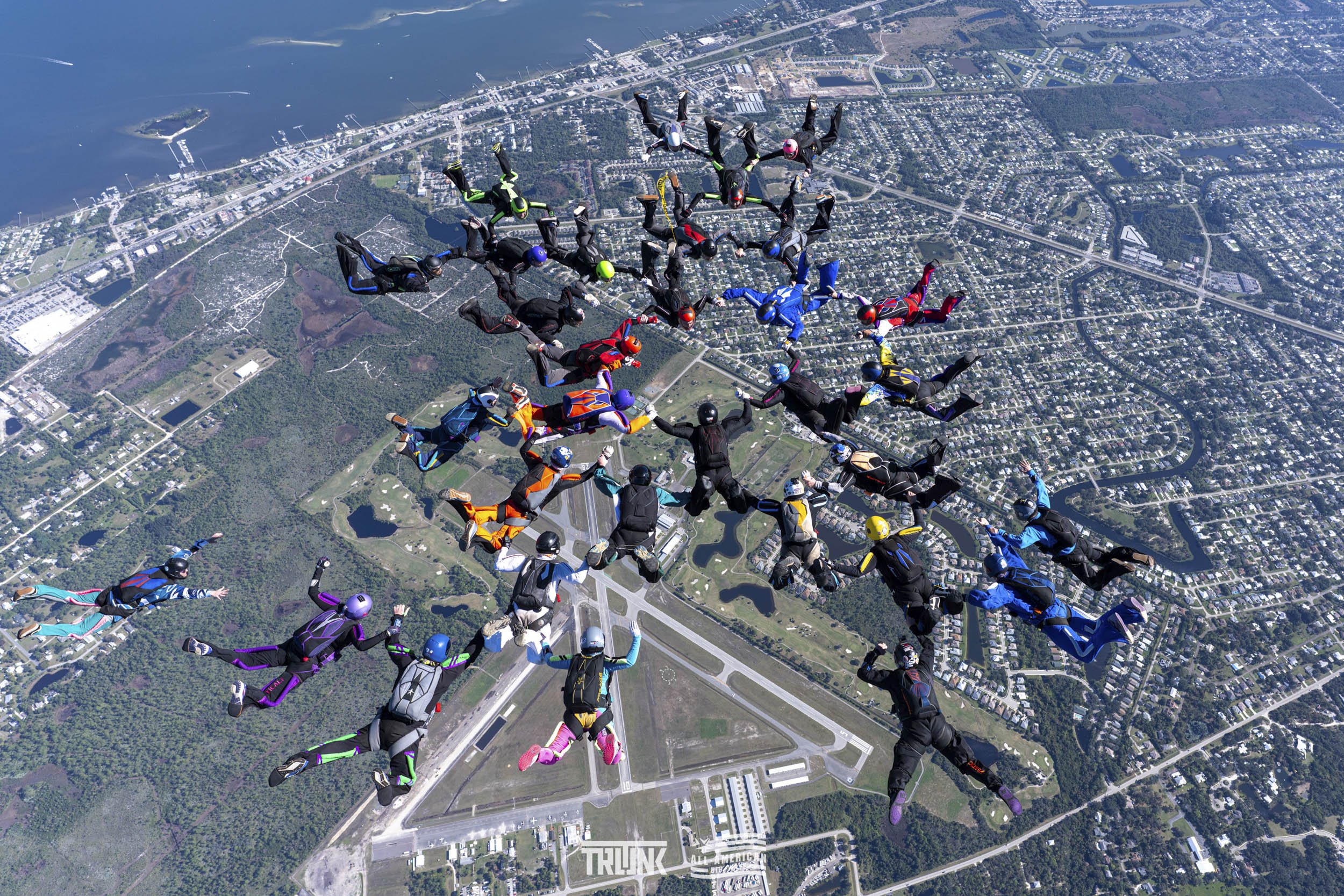Group of skydivers in colorful jumpsuits and helmets mid-air above a cityscape with water, roads, and green areas.