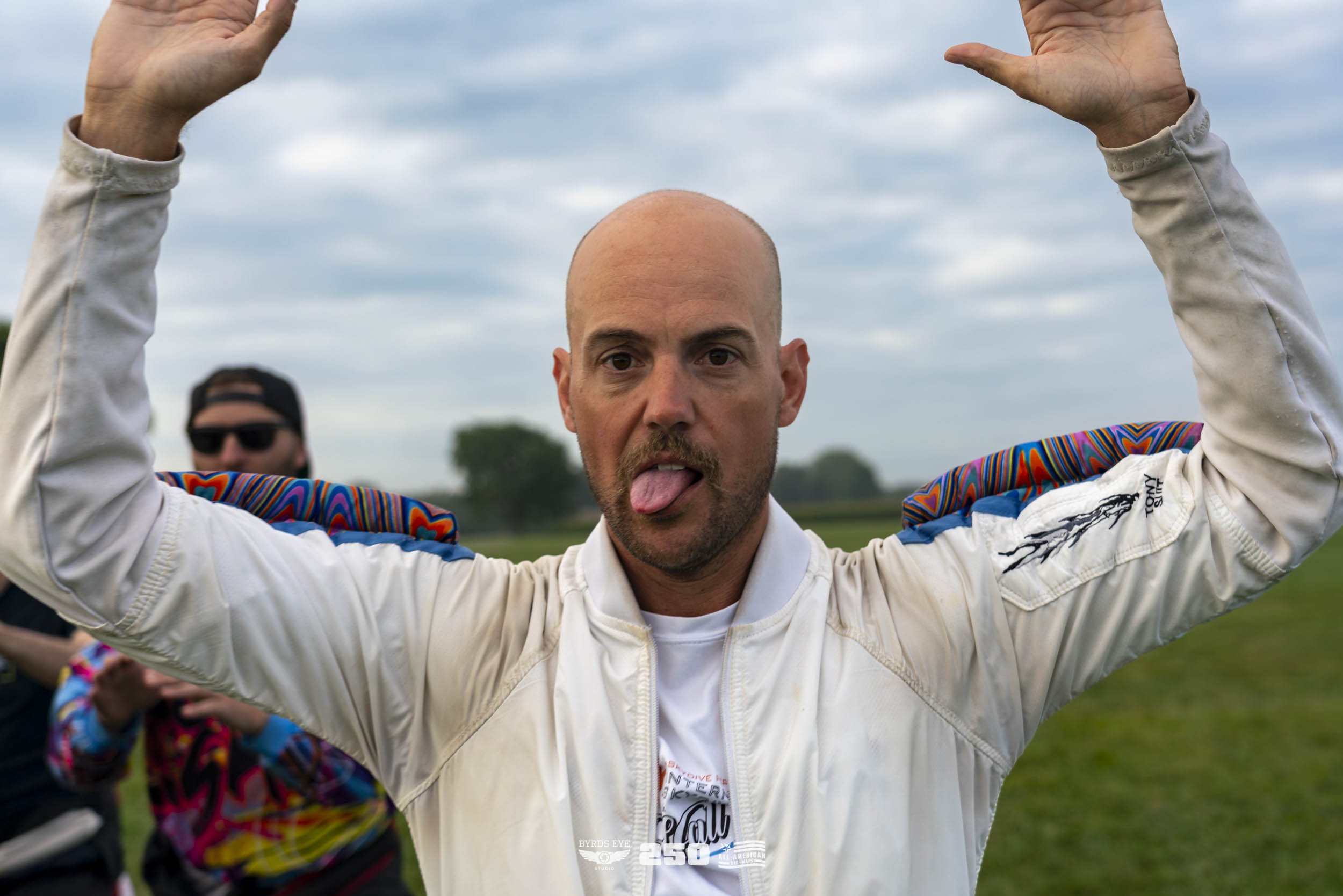 A man with a bald head is standing outdoors in a grassy field, sticking out his tongue and raising both arms above his head, wearing a white jacket with colorful patterned shoulders. There are other people in the background.