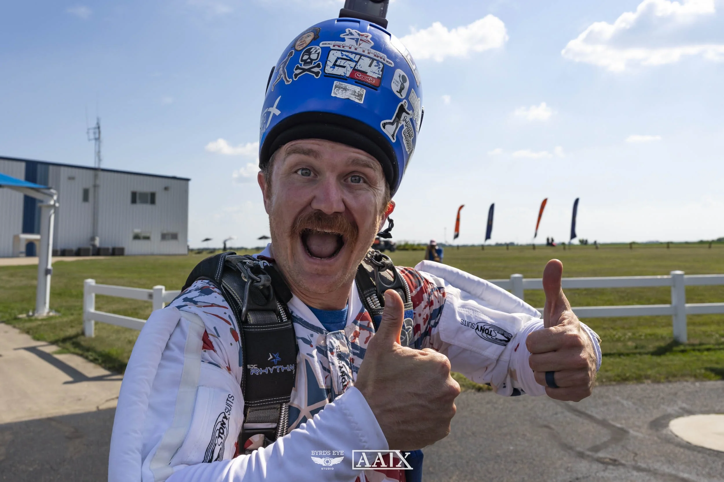A jubilant man wearing a helmet and racing suit giving a thumbs-up at an airfield with flags in the background.