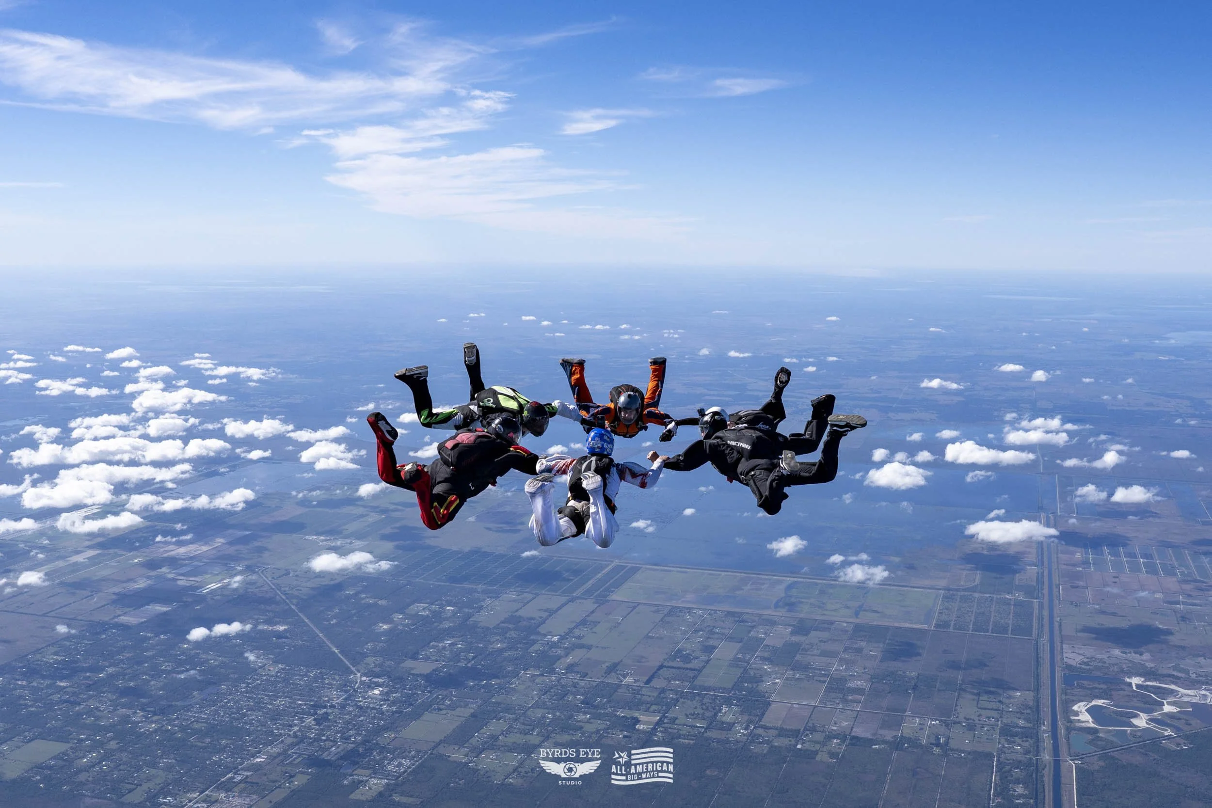 Five skydivers in free fall holding hands forming a circle above the landscape with clouds below.