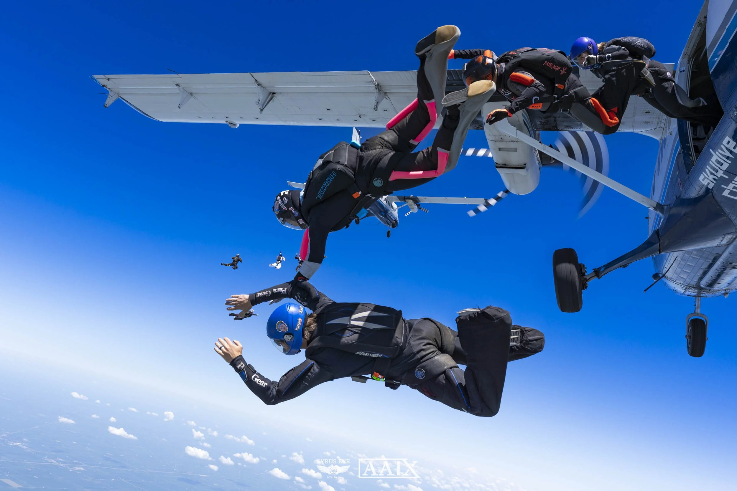 Skydivers exiting a helicopter mid-air, with blue sky and clouds below, preparing for freefall.