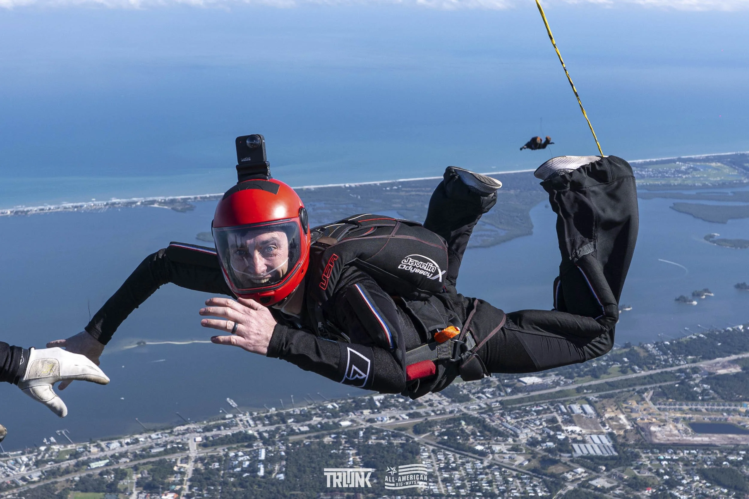 A skydiver in a black jumpsuit and red helmet is mid-air during a jump, with an ocean and a coastal town below.