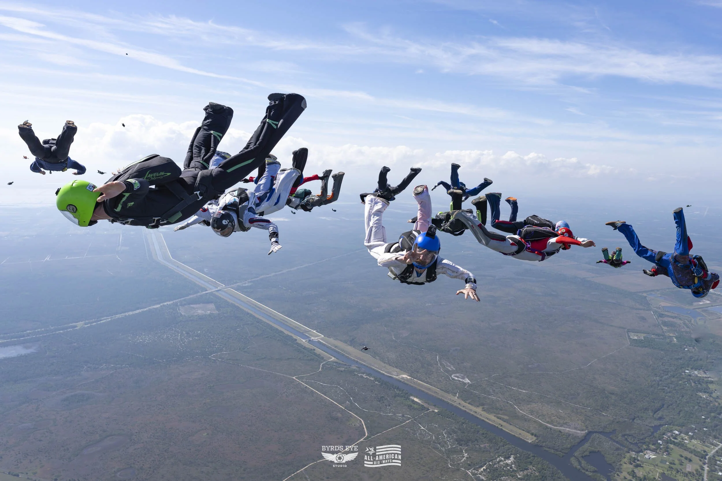 Multiple skydivers in free fall formation above a landscape with roads and fields, under a partly cloudy sky.