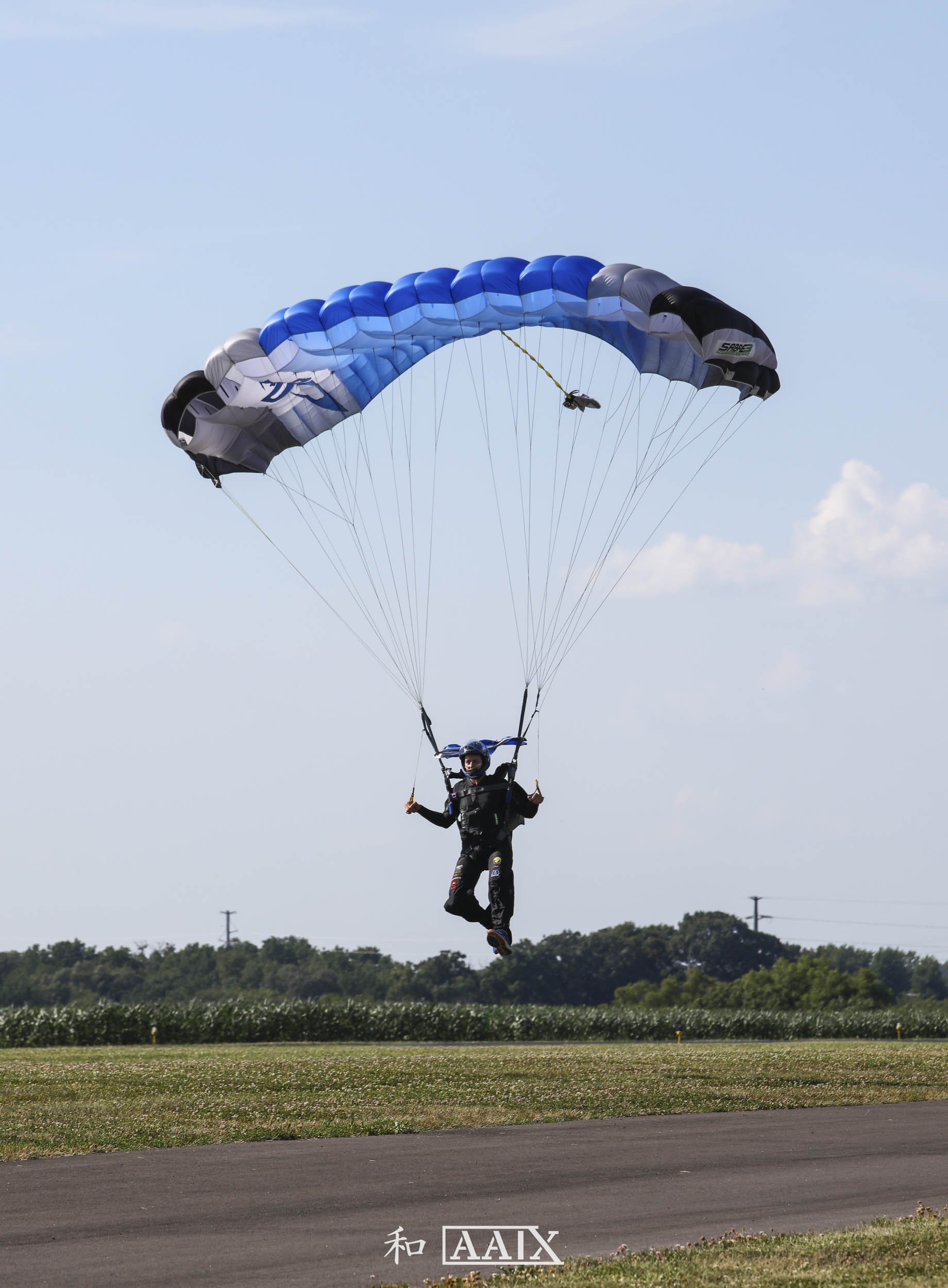 A person in skydiving gear descending with a blue, white, black, and gray parachute over a grassy field and blue sky with clouds.