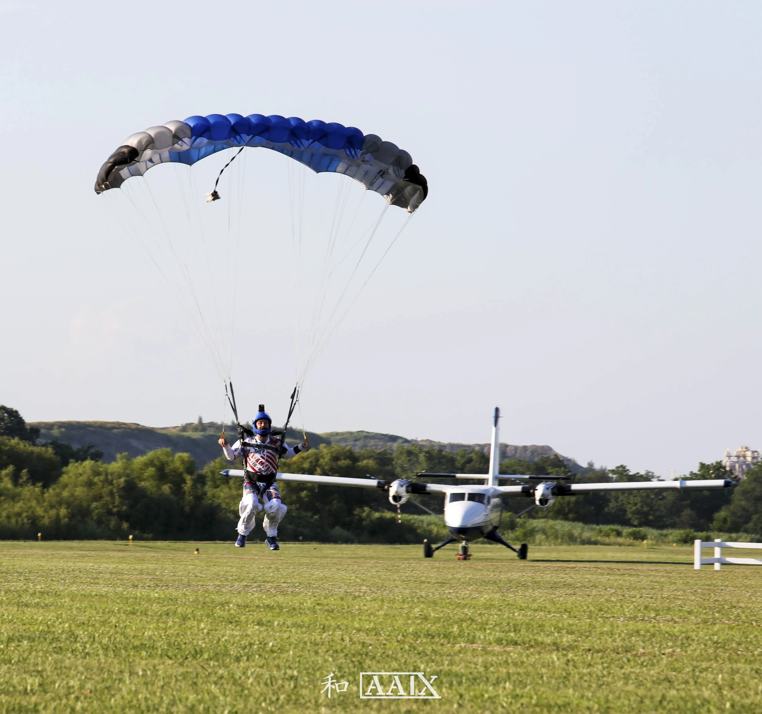 A skydiver descending with a blue and gray parachute near a small airplane on a grassy field, with trees and hills in the background.
