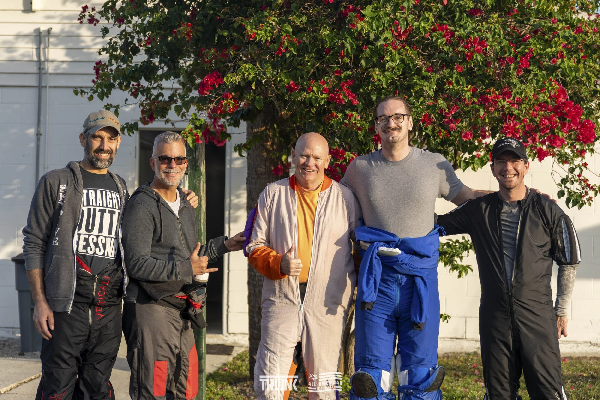 Five men standing outdoors in front of a flowering tree, smiling for the camera.