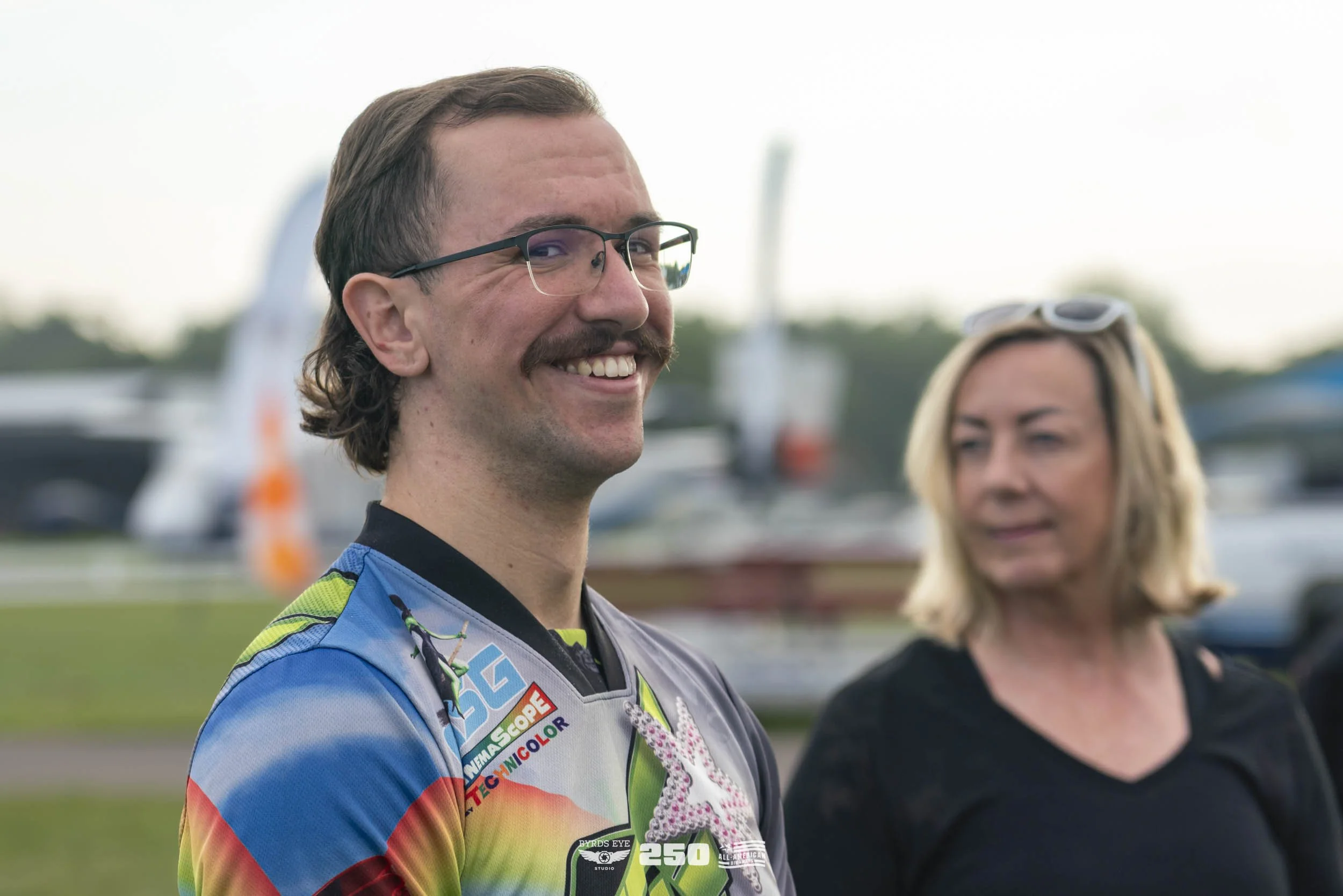 A man with glasses and a beard wearing a colorful sports jersey smiling outdoors, with a woman with blonde hair and sunglasses in the background.