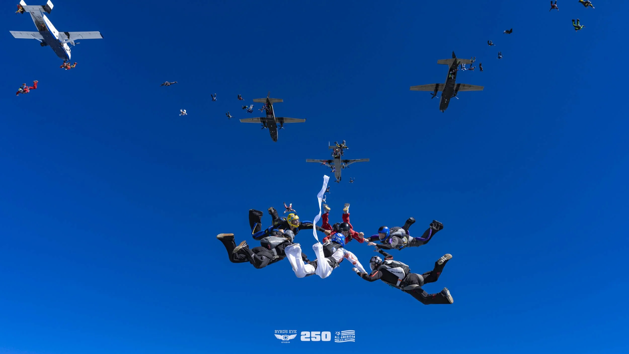 A group of skydivers holding hands in free fall with a clear blue sky and multiple aircraft flying above them.