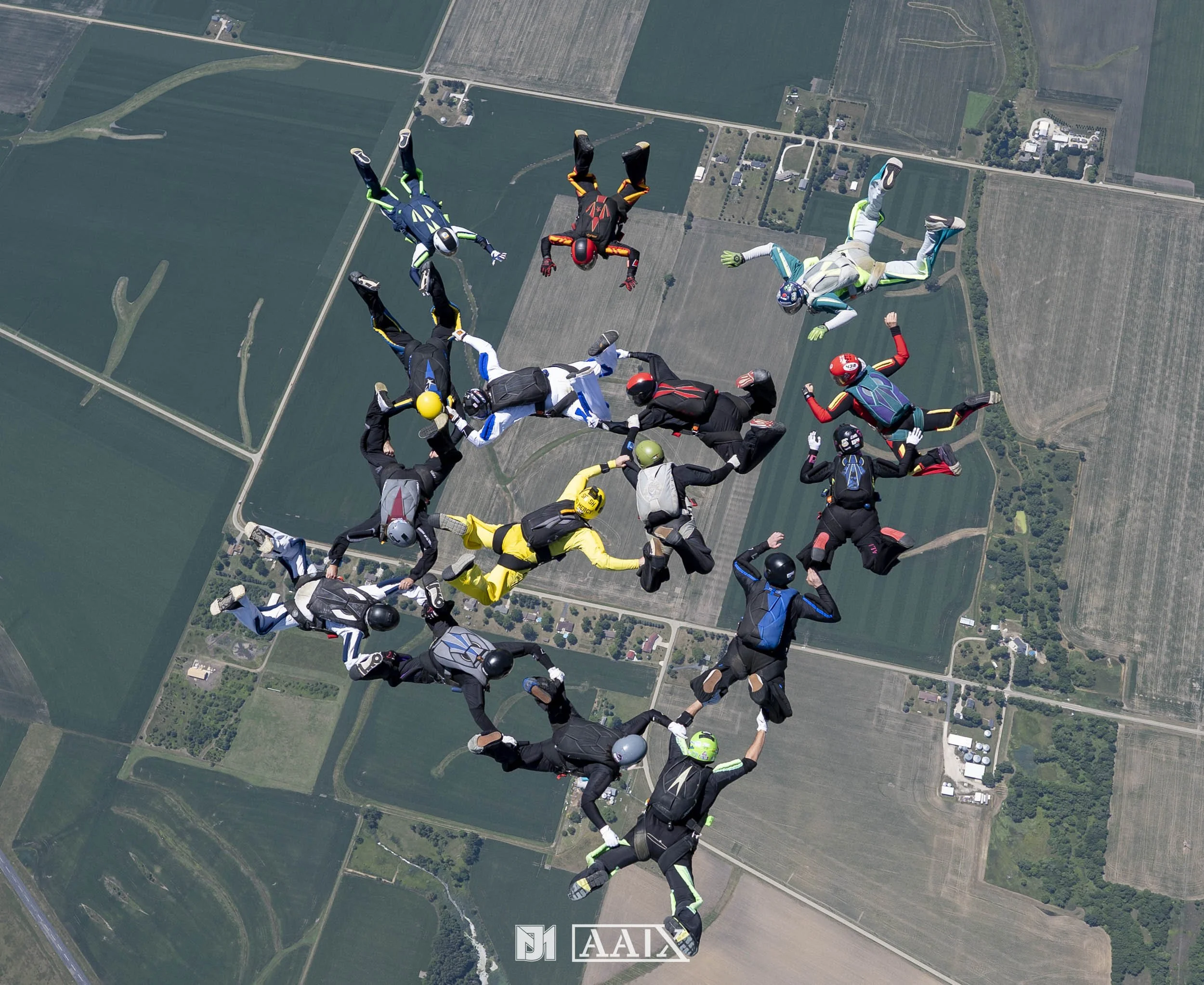 A group of skydivers forming a heart shape mid-air above a patchwork of fields and rural landscape.