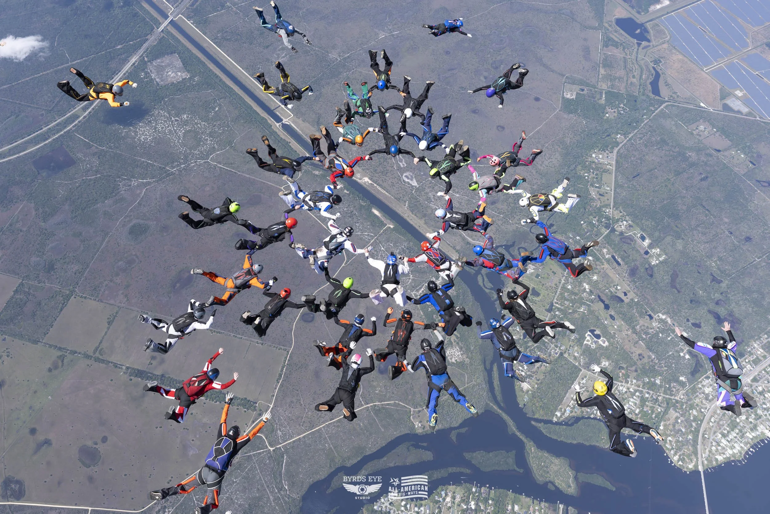 Numerous skydivers in colorful suits and helmets are free-falling together above a landscape with rivers, roads, and fields.