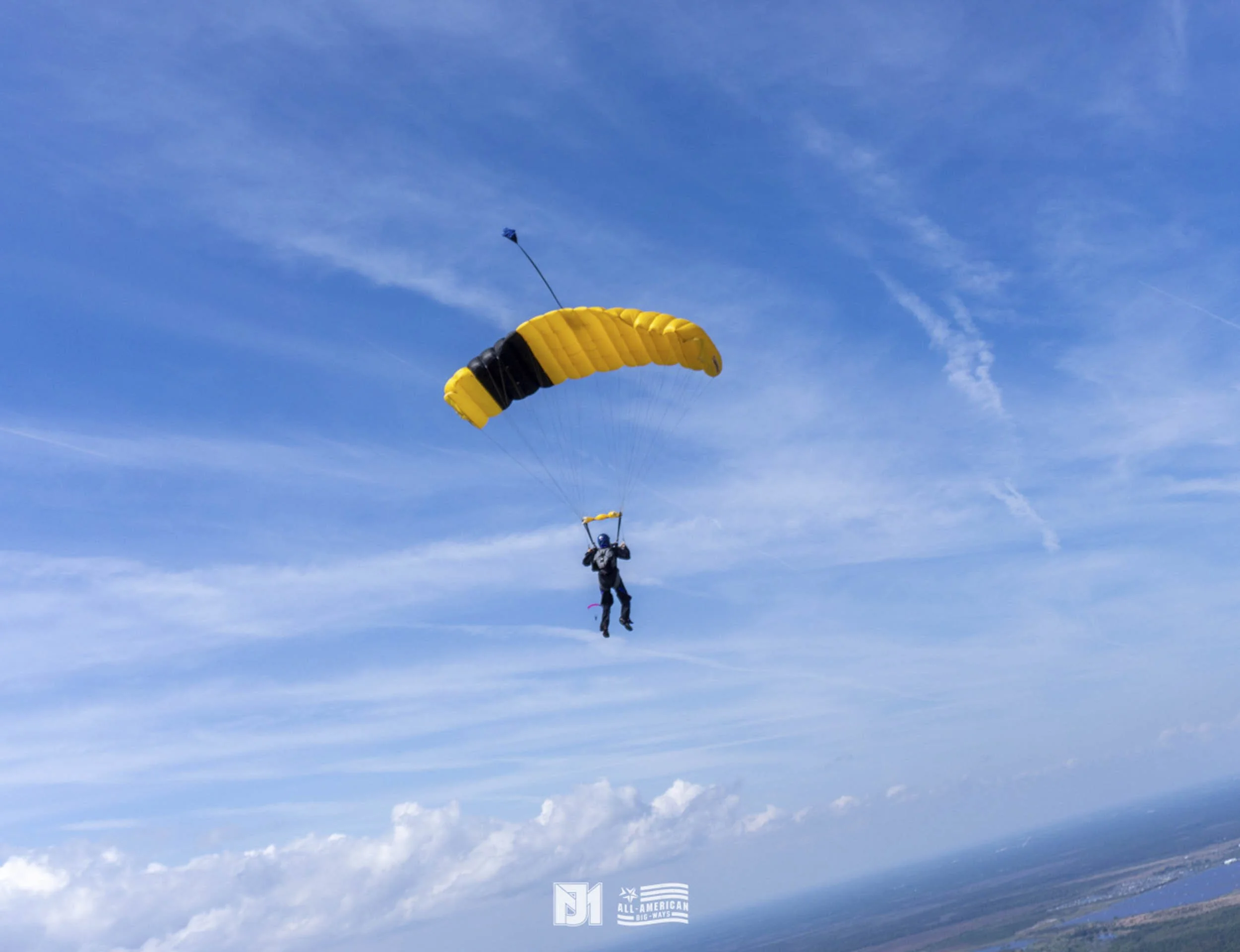A person skydiving with a yellow and black parachute against a blue sky with scattered clouds.