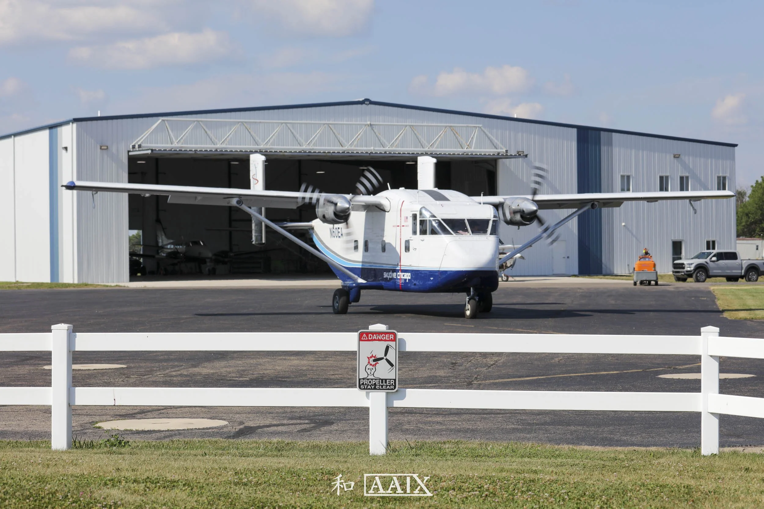 Small white and blue amphibious aircraft parked on tarmac outside a hangar with open doors, behind a white fence with a danger propeller sign.