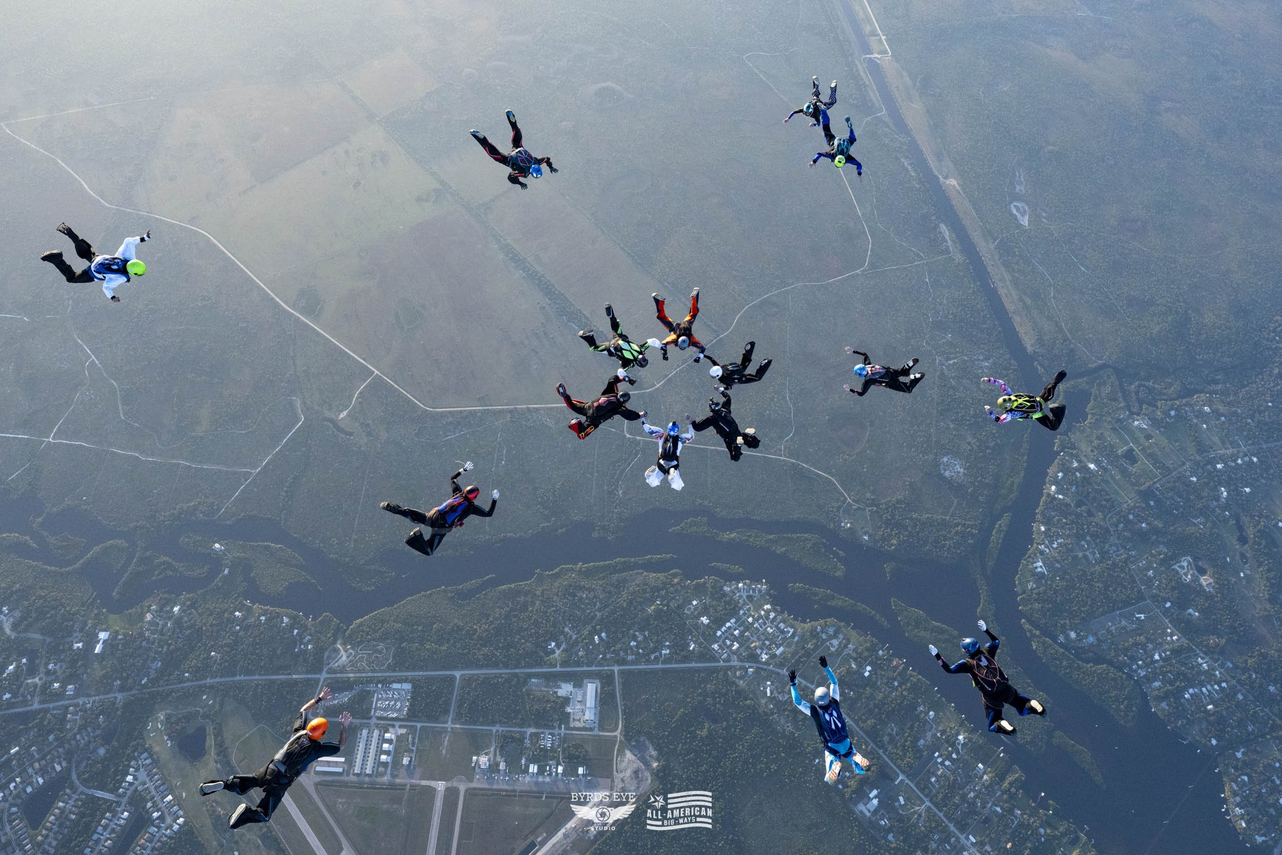 Group of skydivers in free fall formation over a landscape with lakes, forests, and roads.