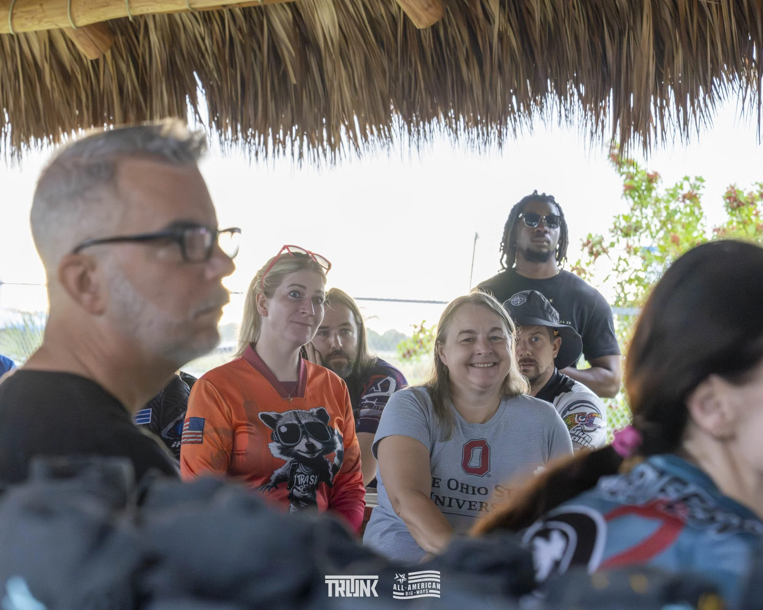 A group of seven diverse people sitting under a thatched roof, some looking toward the camera, some looking elsewhere, outdoors on a sunny day.